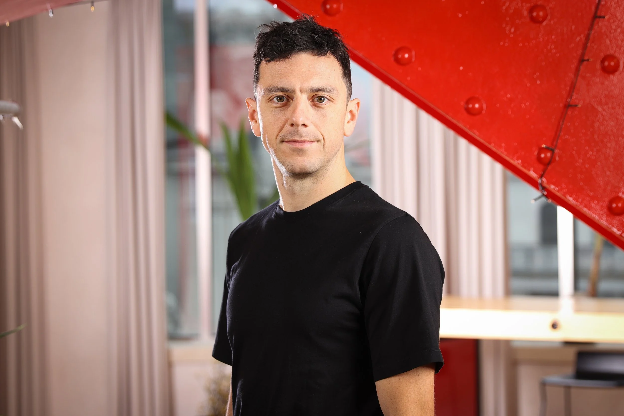 A man with short dark hair wearing a black t-shirt standing indoors near a red grand piano, with large windows and beige curtains in the background.