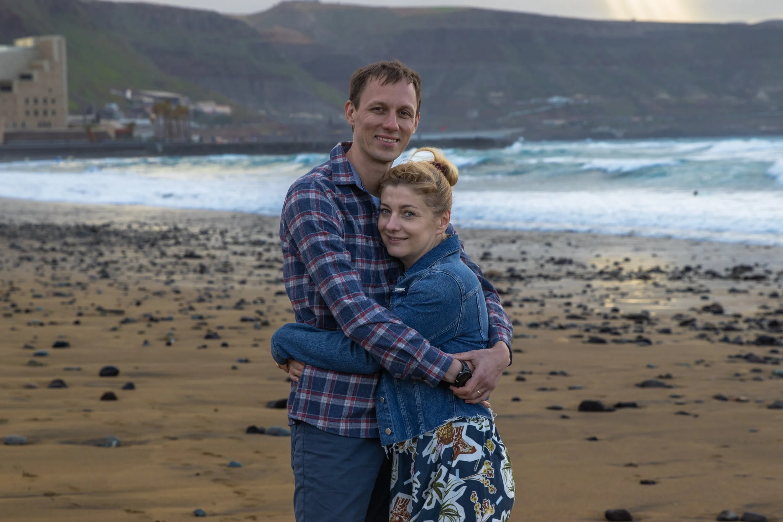 A couple hugging on a beach, with the waves and cliffs in the background.