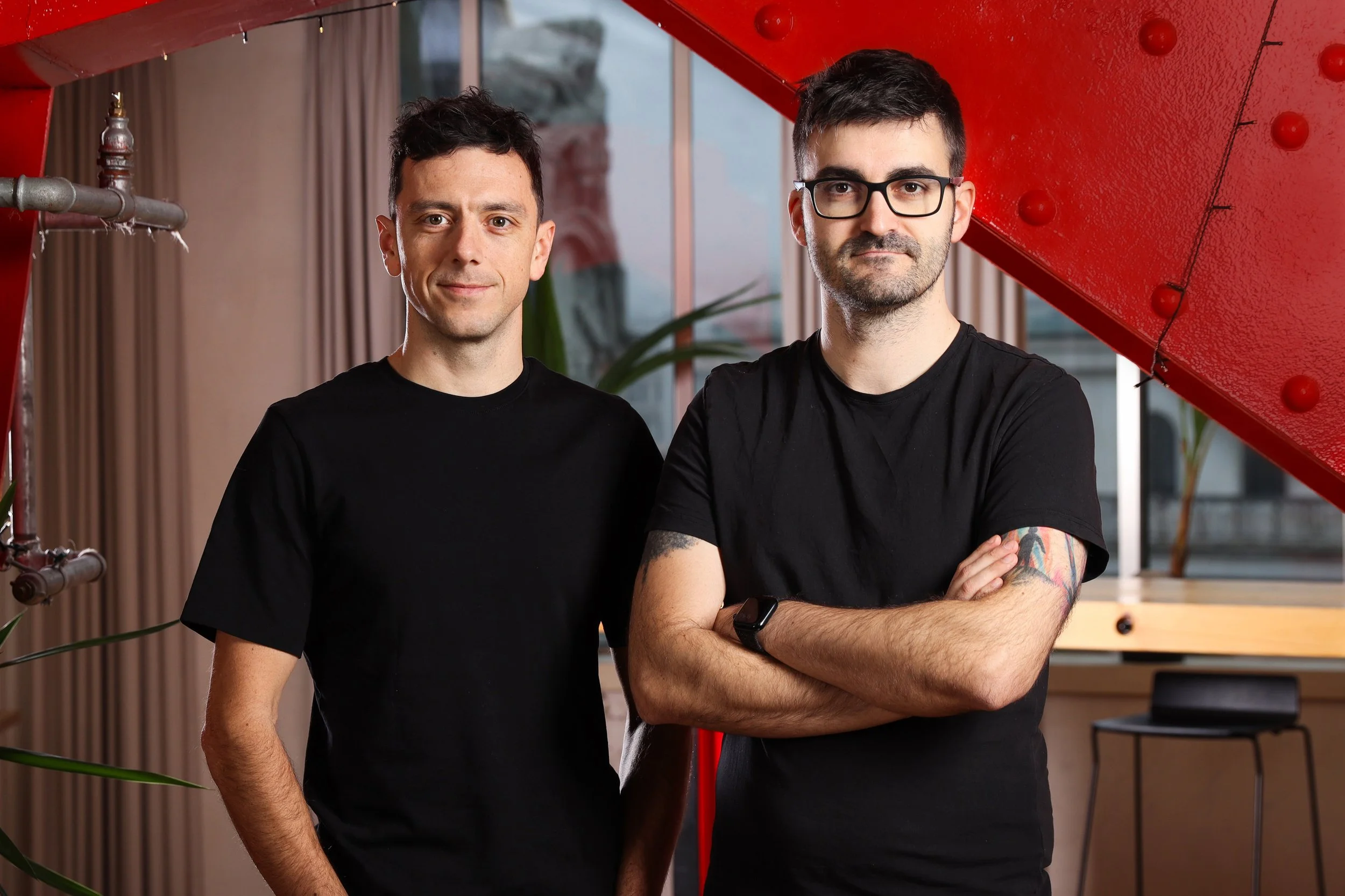 Two men standing indoors in front of a window and a red staircase, both wearing black t-shirts. The man on the right has glasses, a beard, a tattoo on his left arm, and is crossing his arms. The man on the left has short dark hair and is smiling.
