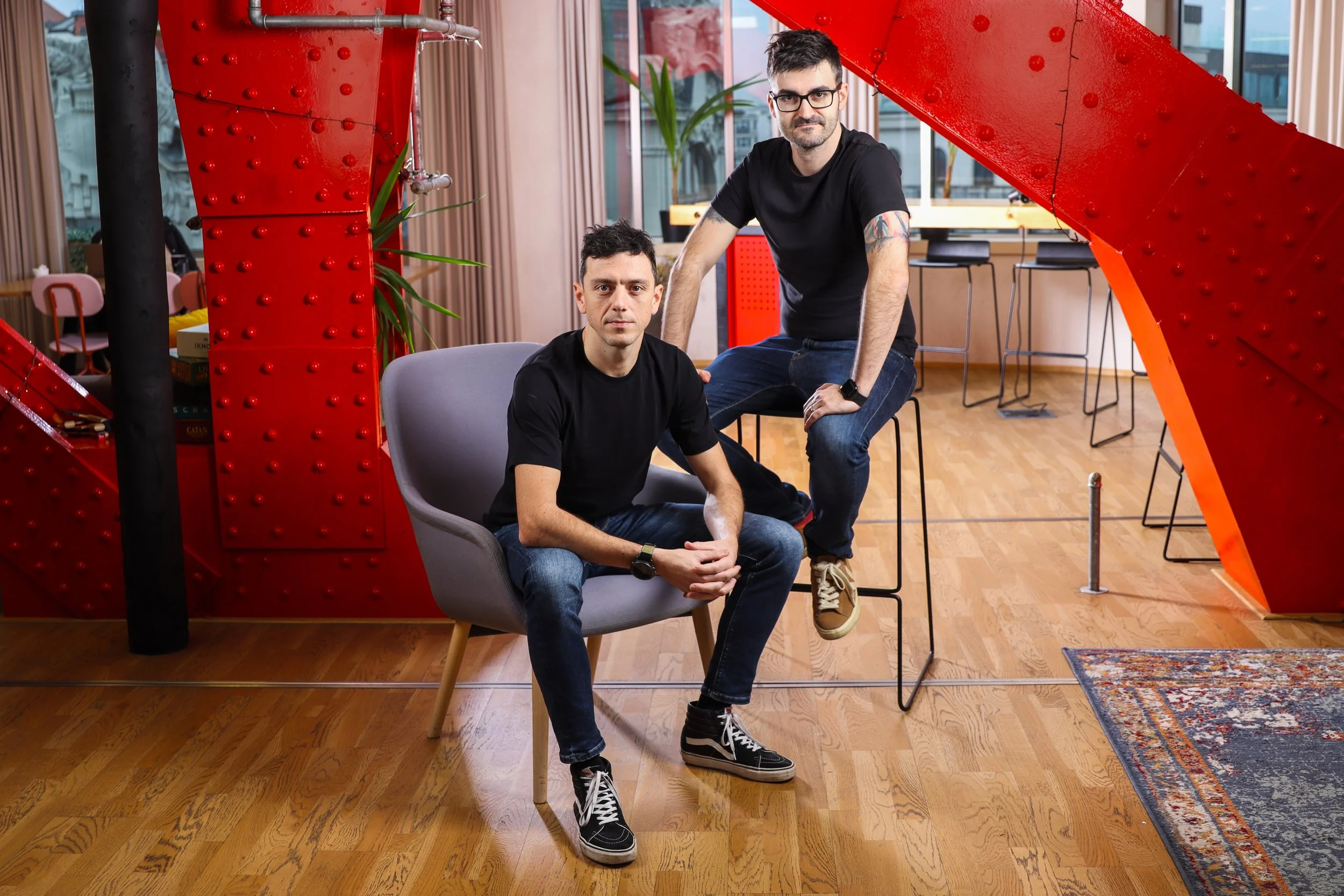 Two young men with dark hair and casual black t-shirts pose in an indoor space with large windows, wooden floors, and modern furniture. One is sitting in a gray chair, and the other is leaning on a barstool.