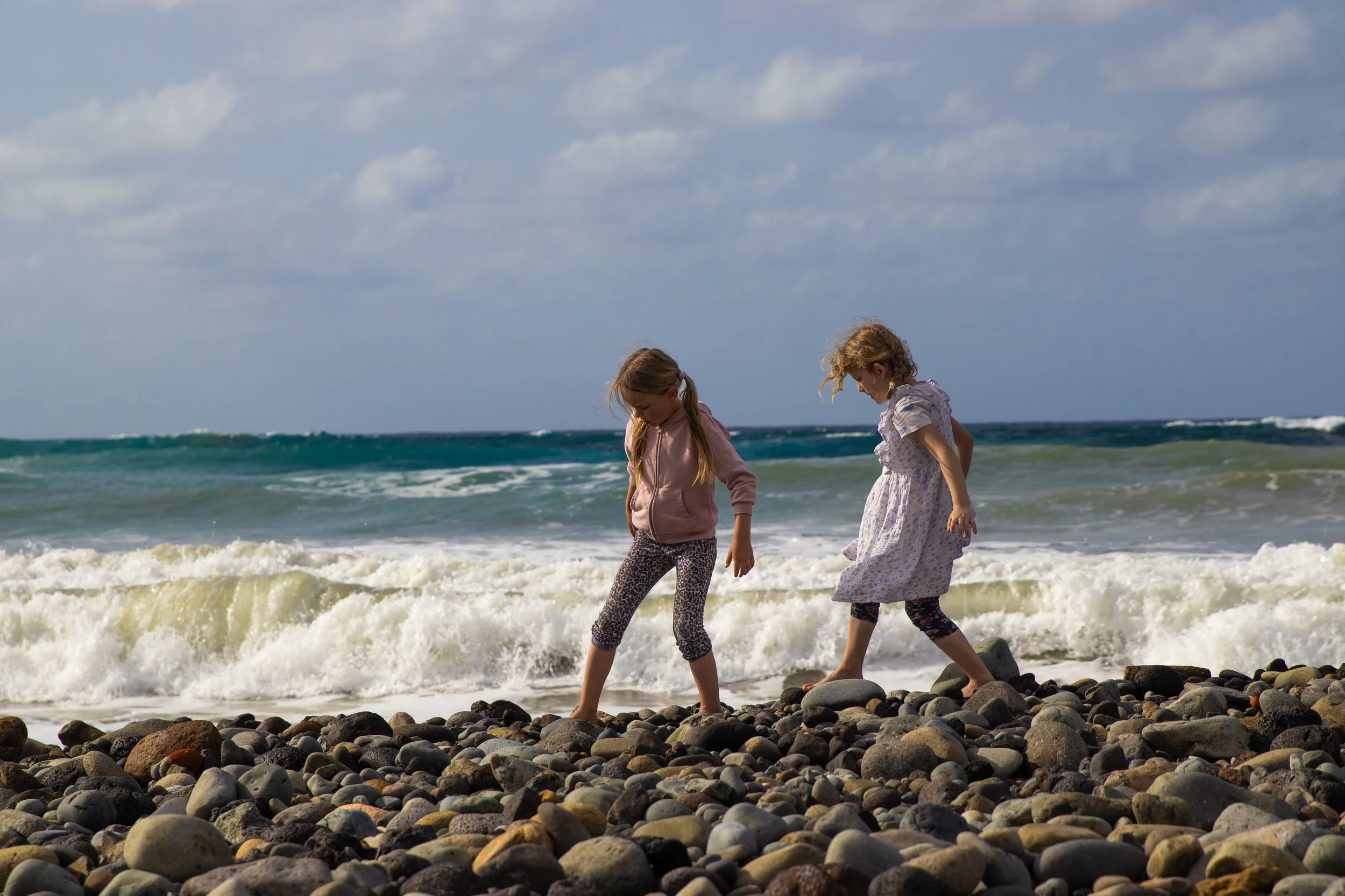 Two young girls walking barefoot on a rocky beach by the ocean, with waves crashing behind them and a partly cloudy sky above.