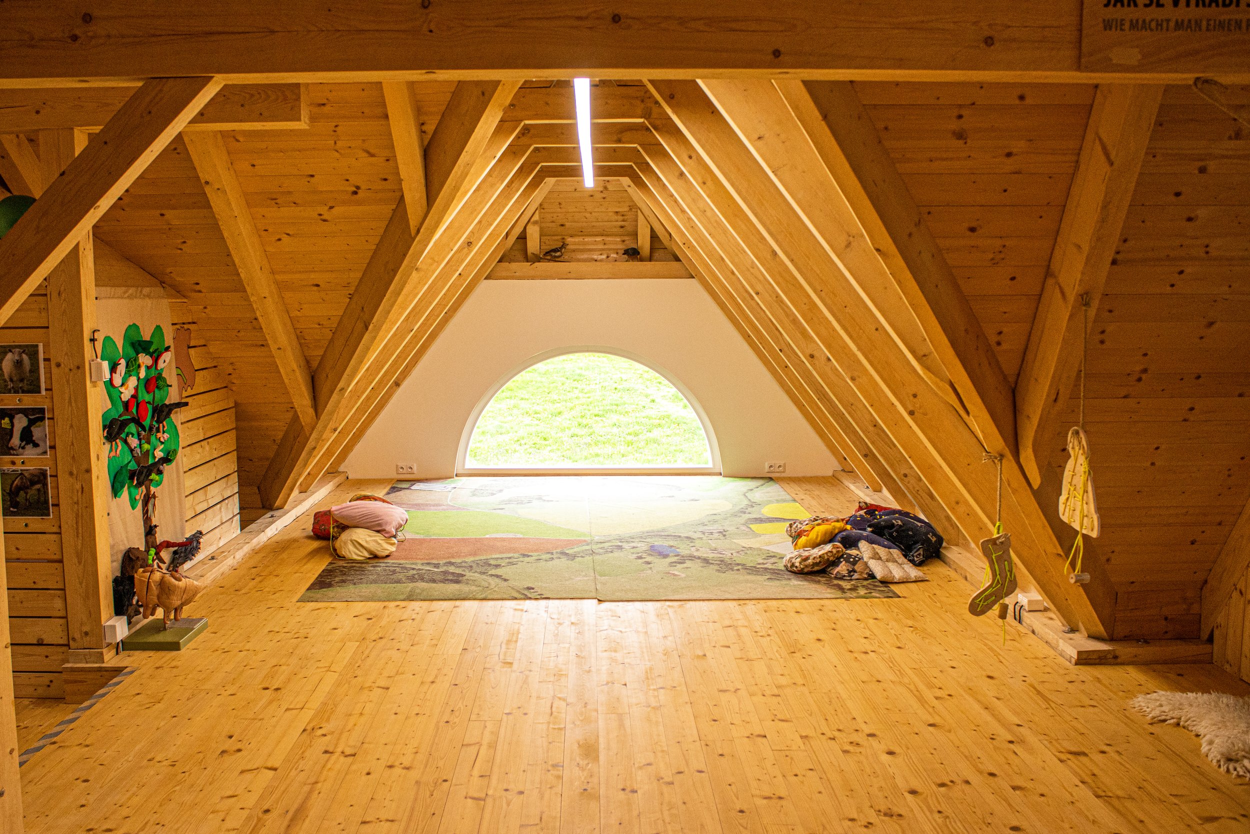 Interior of an attic with wooden beams and flooring, a large arched window at the far end, and colorful pillows and blankets on the floor.