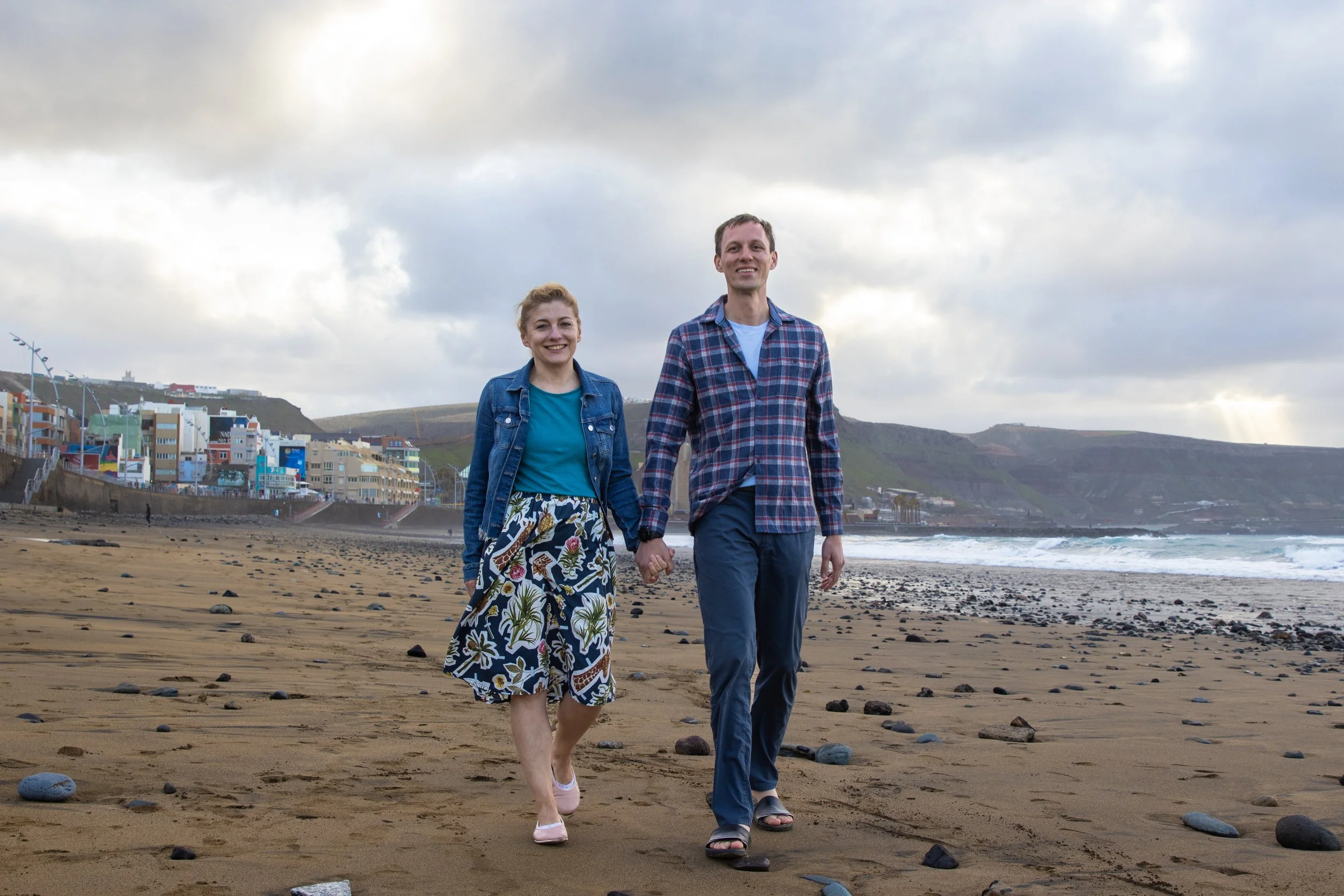 A couple walking hand in hand on a beach with colorful buildings on a hillside in the background and cloudy sky.
