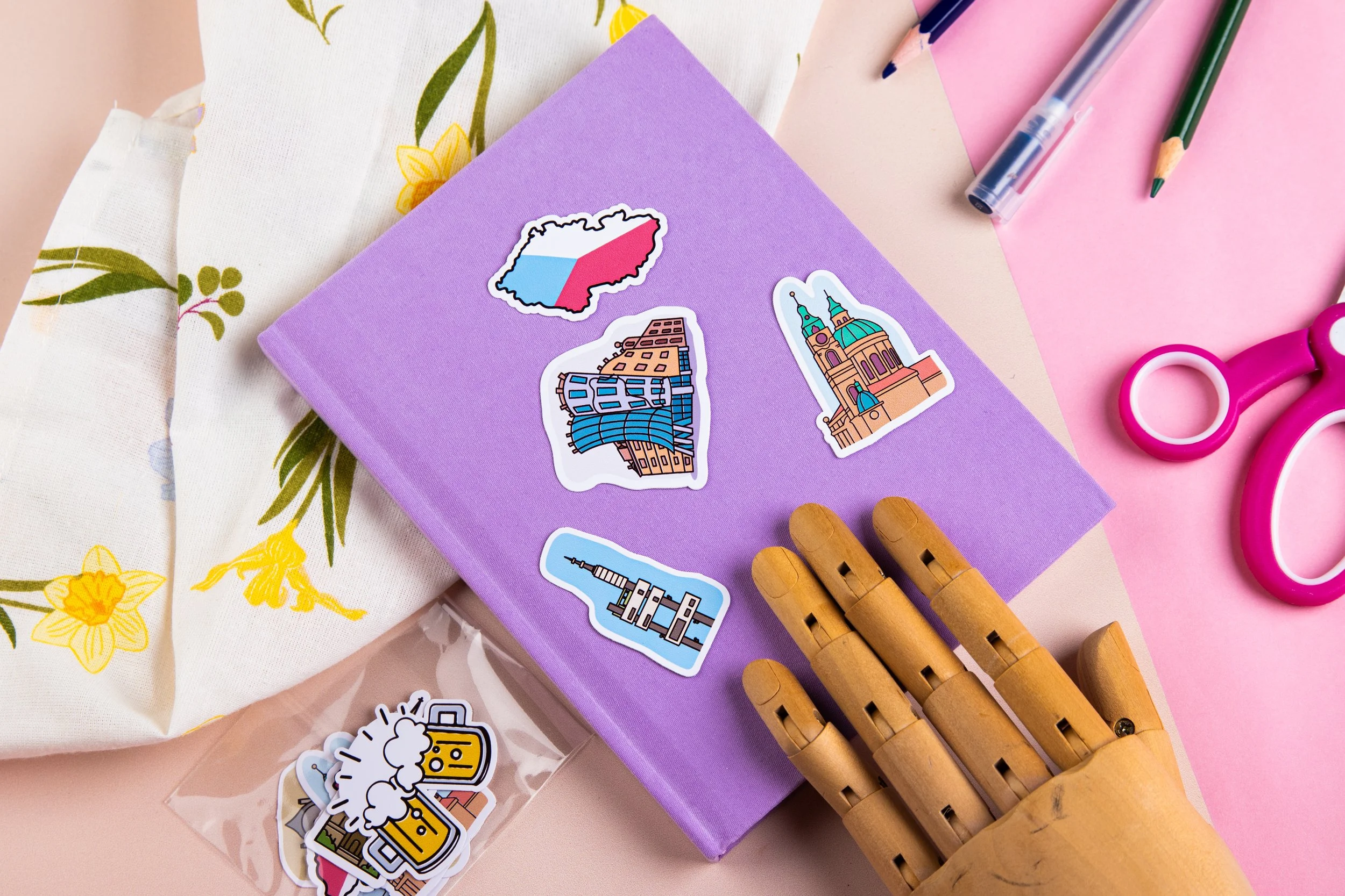 A purple notebook on a pink and cream surface with several colorful travel sticker decals, a wooden robotic hand, pink scissors, markers, and pens, alongside a floral-printed fabric.