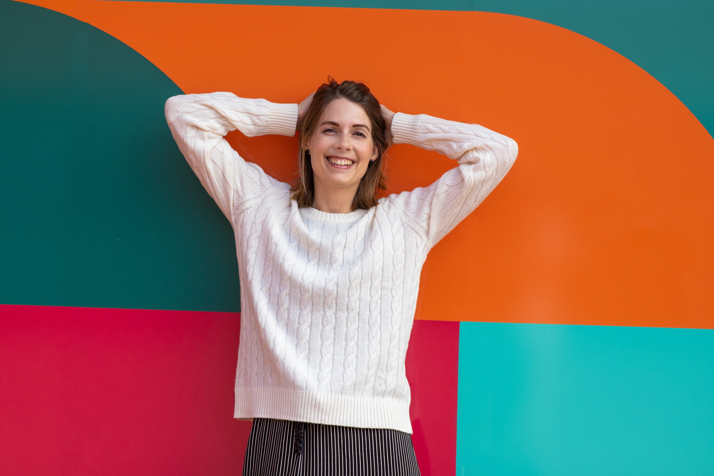 A young woman with shoulder-length brown hair, smiling and standing with hands behind her head in front of a colorful geometric wall background.