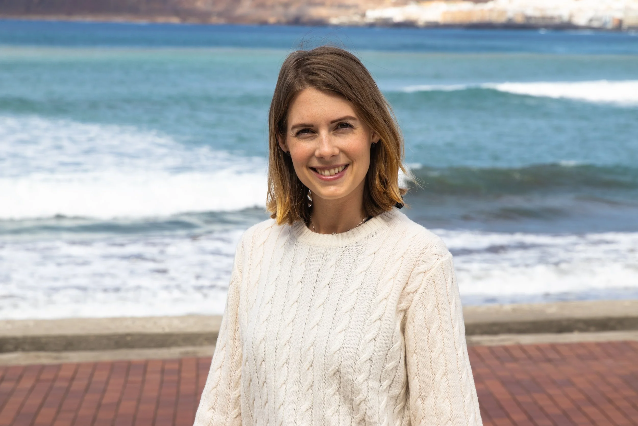 A smiling young woman with shoulder-length brown hair wearing a white knitted sweater standing on a brick-paved pathway near the ocean, with waves and a distant shoreline in the background.