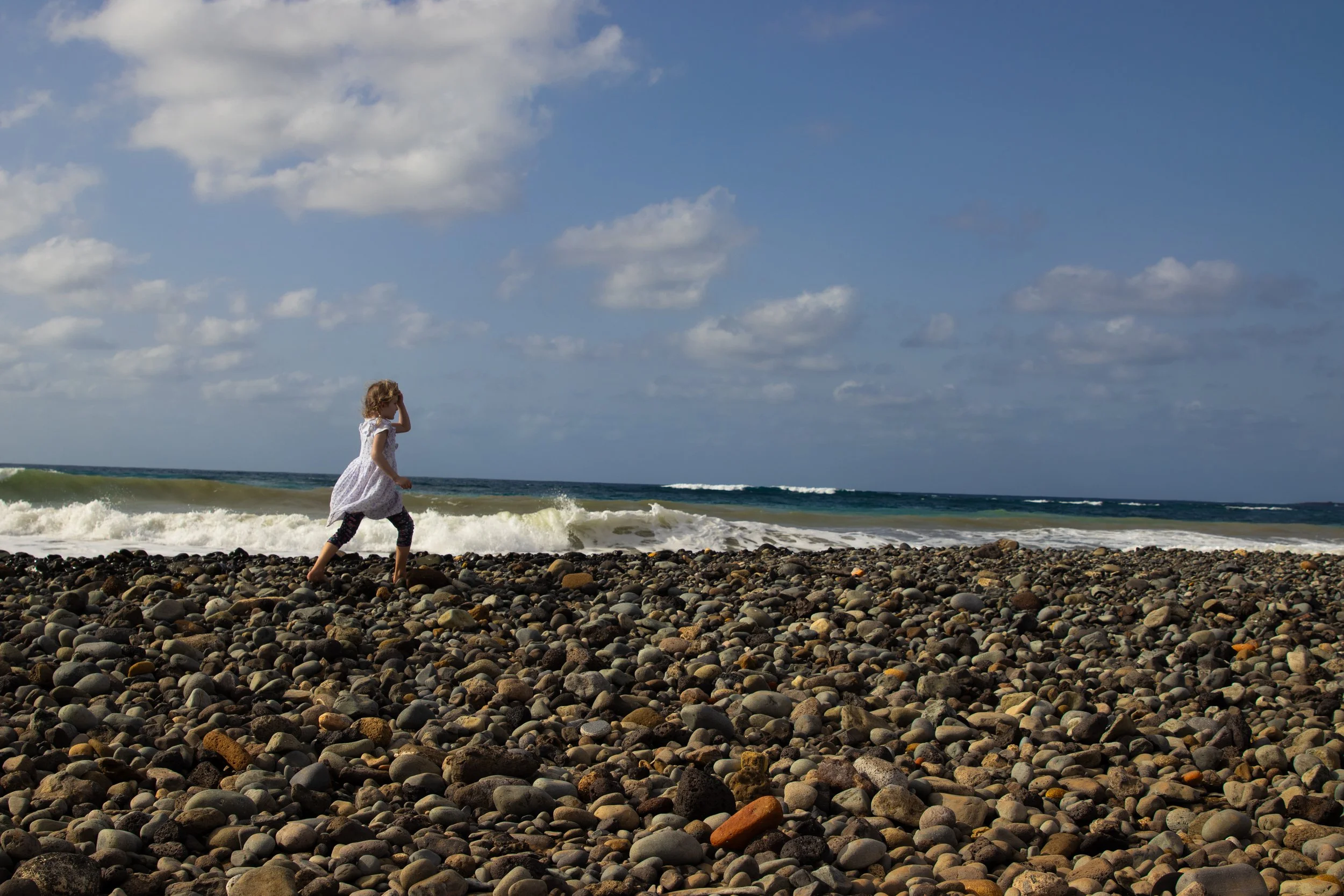 A young girl in a white dress and leggings standing on a pebbled beach, looking toward the ocean with waves crashing and a partly cloudy sky overhead.