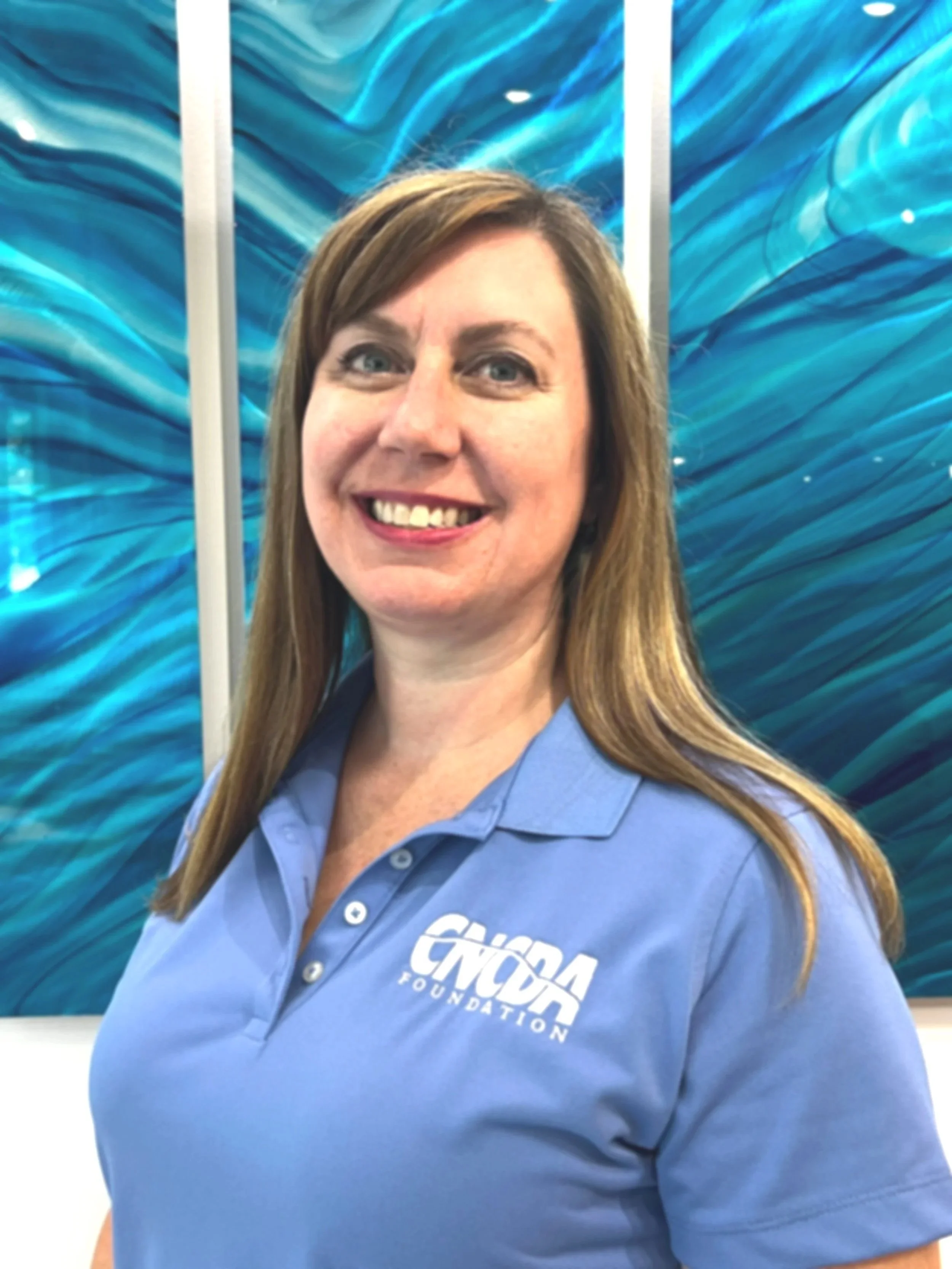 A woman wearing a blue polo shirt with the logo 'CNCA FOUNDATION' standing in front of abstract blue art.