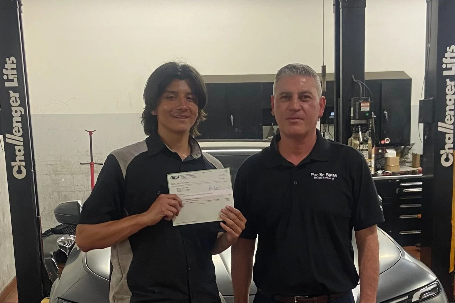 Two men standing in a garage, one holding a certificate, the other wearing a black shirt with 'Pacific BMW' logo, in front of a silver BMW car.