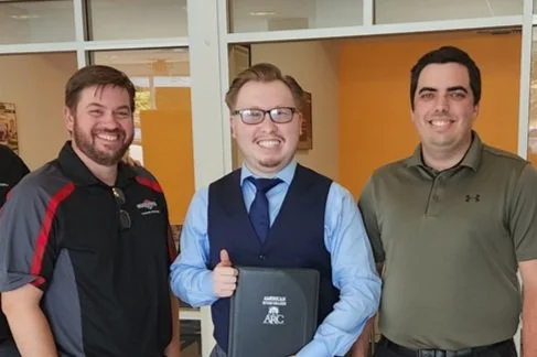 Three men pose for a photo indoors, one middle-aged man in a blue shirt, glasses, and tie holds a folder and gives a thumbs-up, flanked by two younger men in casual attire.