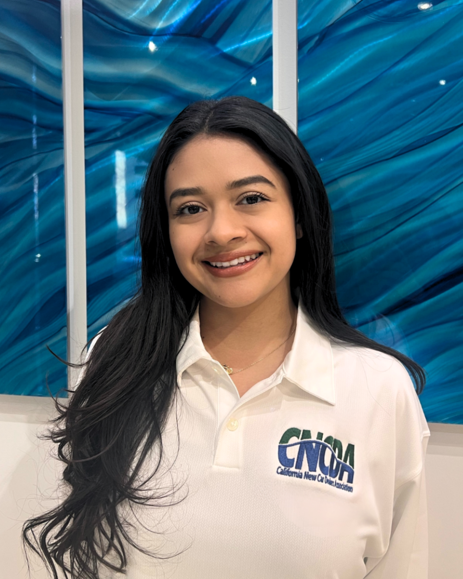 A young woman with long brown hair, wearing a white shirt standing in front of a vibrant, abstract blue and teal artwork.