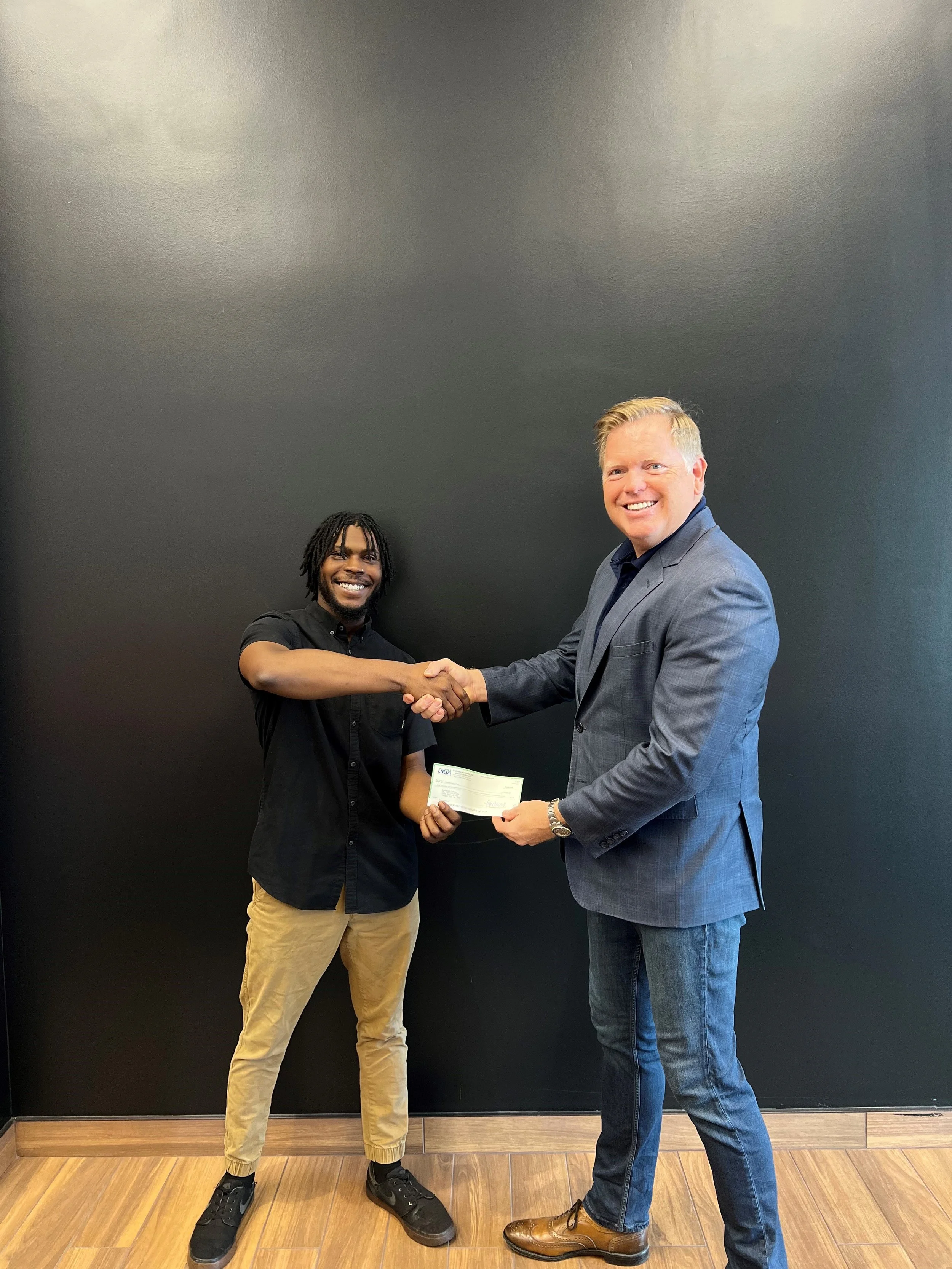 Two men shaking hands; the man on the left holds a check, both smiling, in front of a black wall.