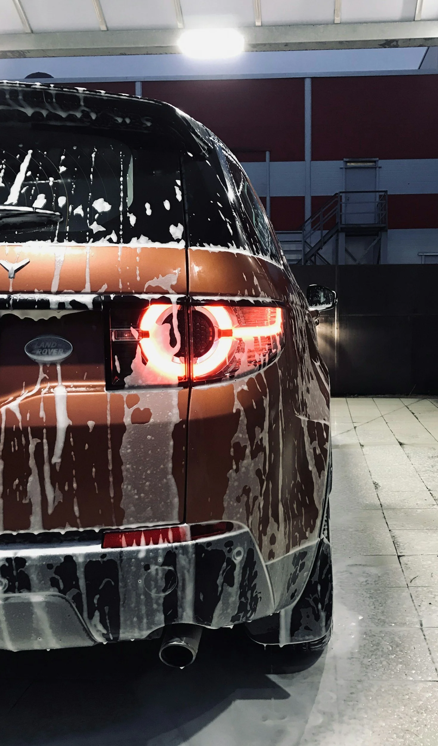 The back of a Land Rover Discovery SUV covered in soap suds, at a car wash. The vehicle's taillights are on, and the setting appears to be an indoor or covered wash station with concrete floor and red and gray wall panels in the background.