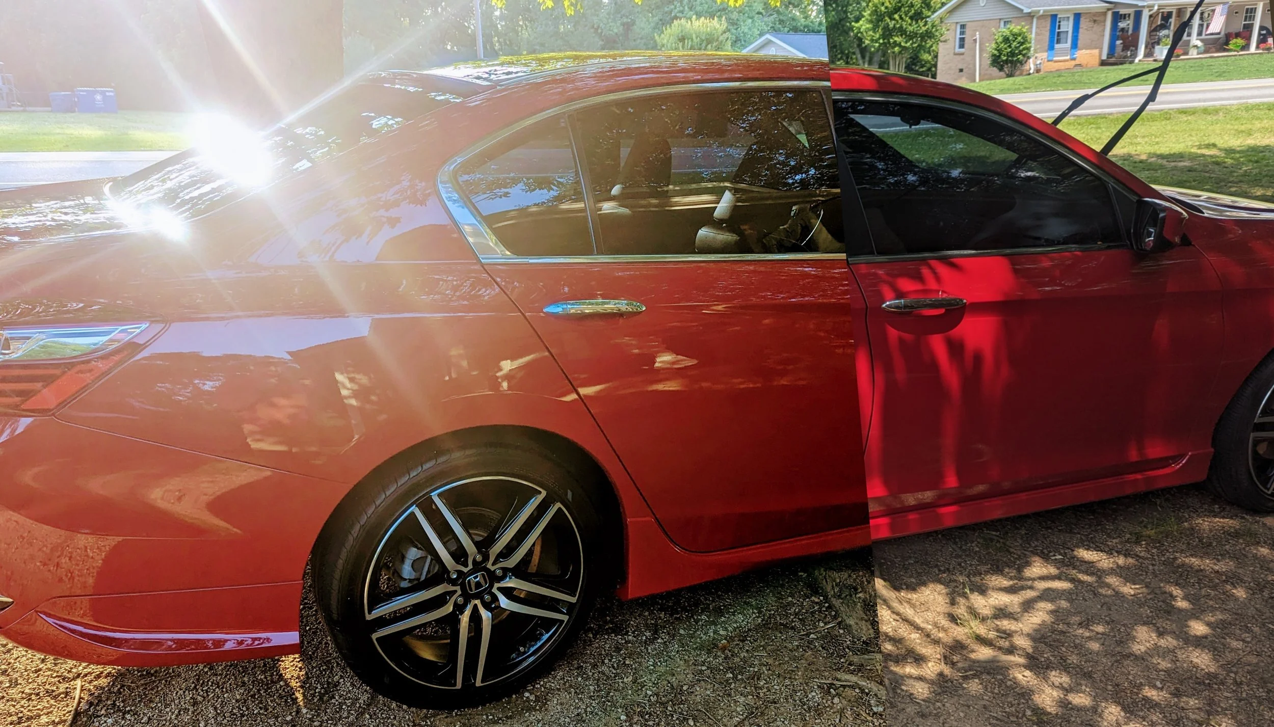 A red sedan parked outdoors with the left half in sunlight and the right half in shade. The car has black alloy wheels and tinted windows, with houses and trees in the background.
