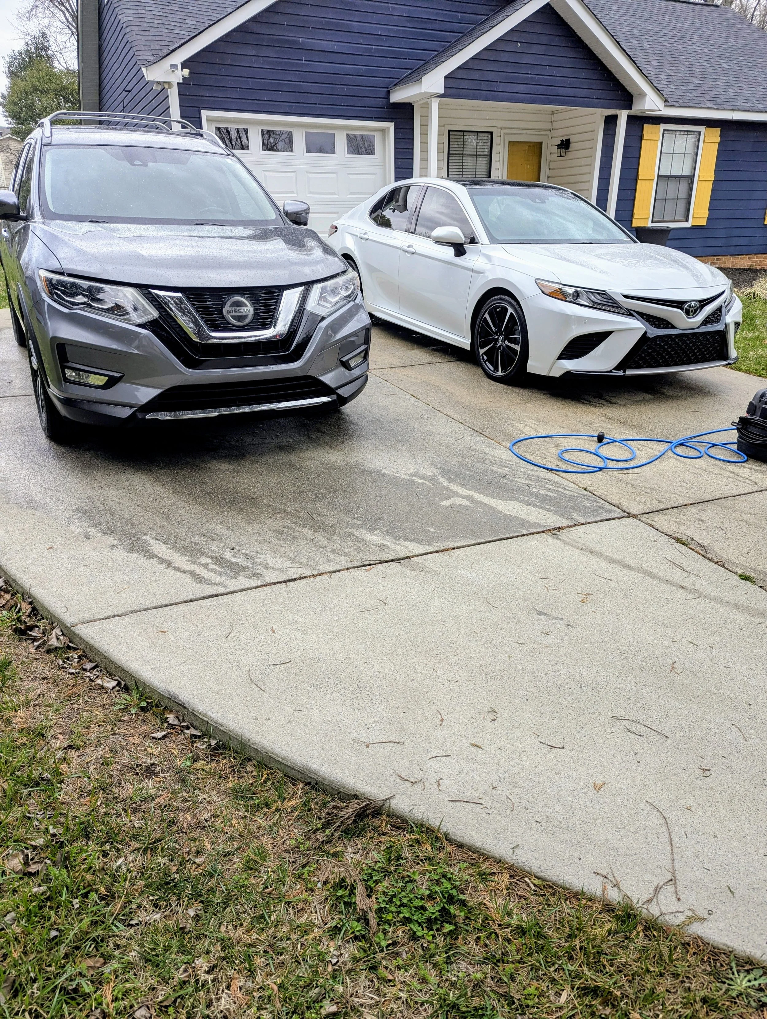 A gray Nissan SUV and a white Toyota sedan parked on a concrete driveway in front of a blue house with yellow window shutters.