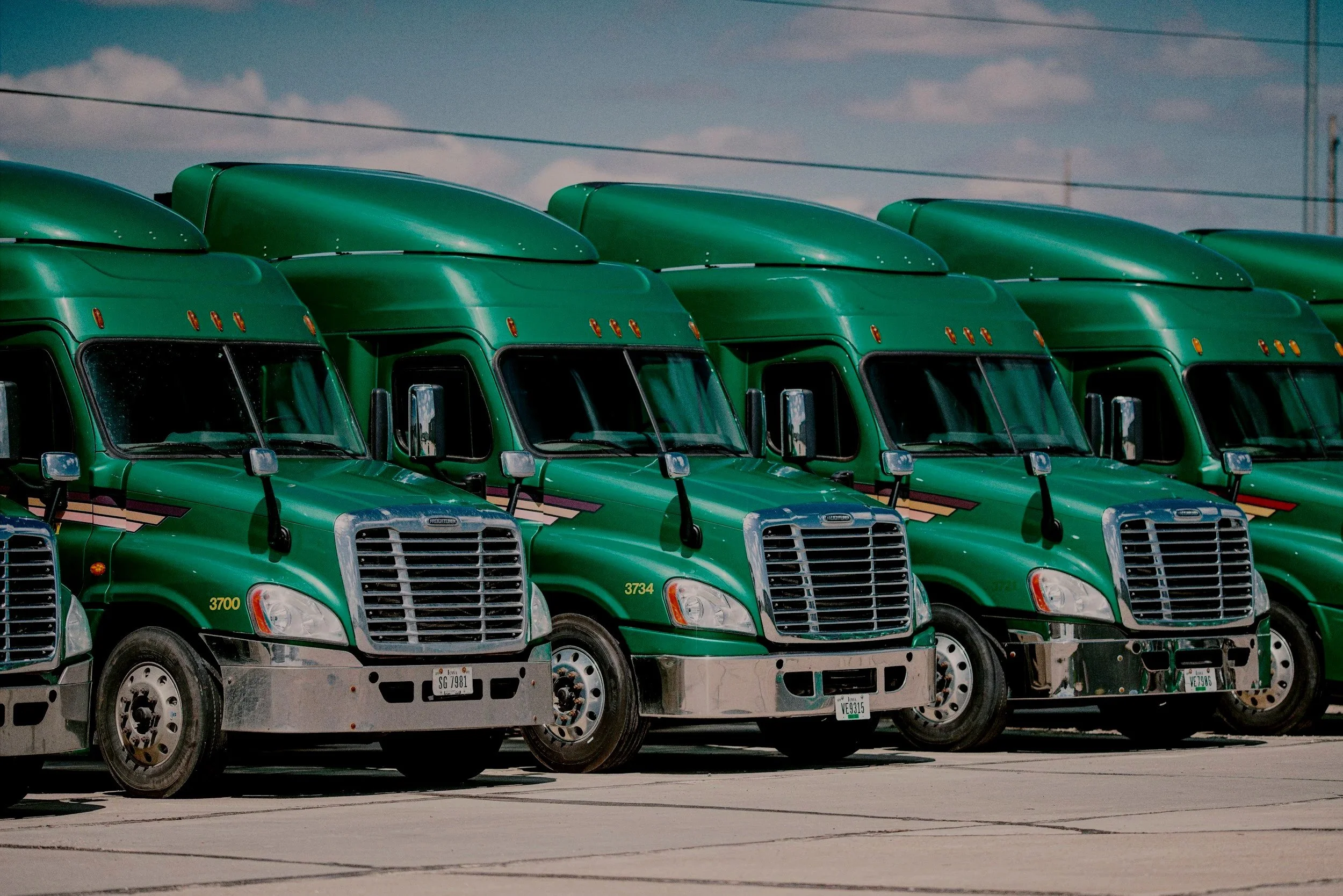A row of green semi-trucks parked side by side in an outdoor lot under a partly cloudy sky.
