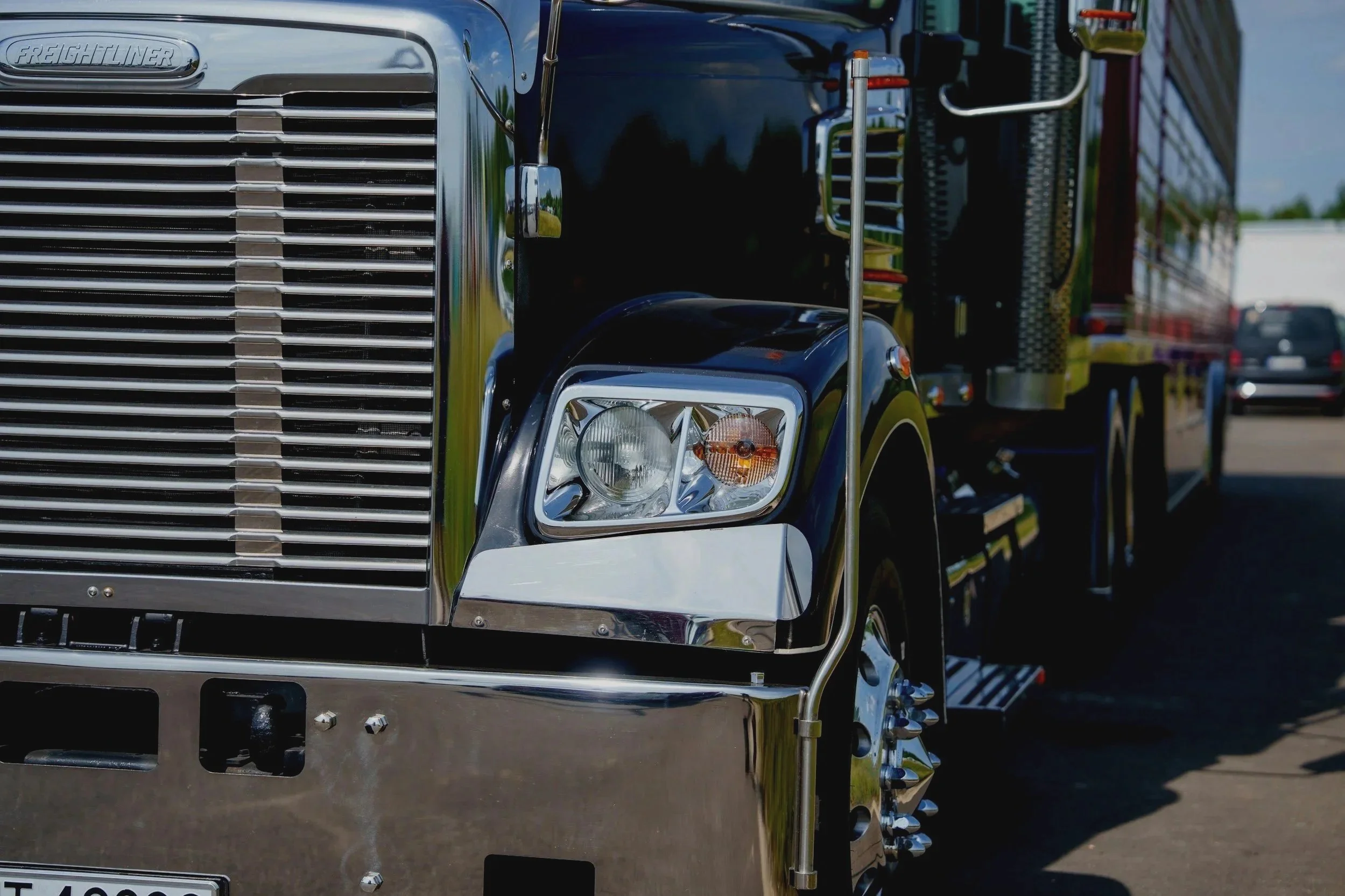 Close-up of the front part of a large black freightliner truck, focusing on the shiny chrome grille, headlight, and side mirrors, with a reflective surface showing a grassy area and blue sky.