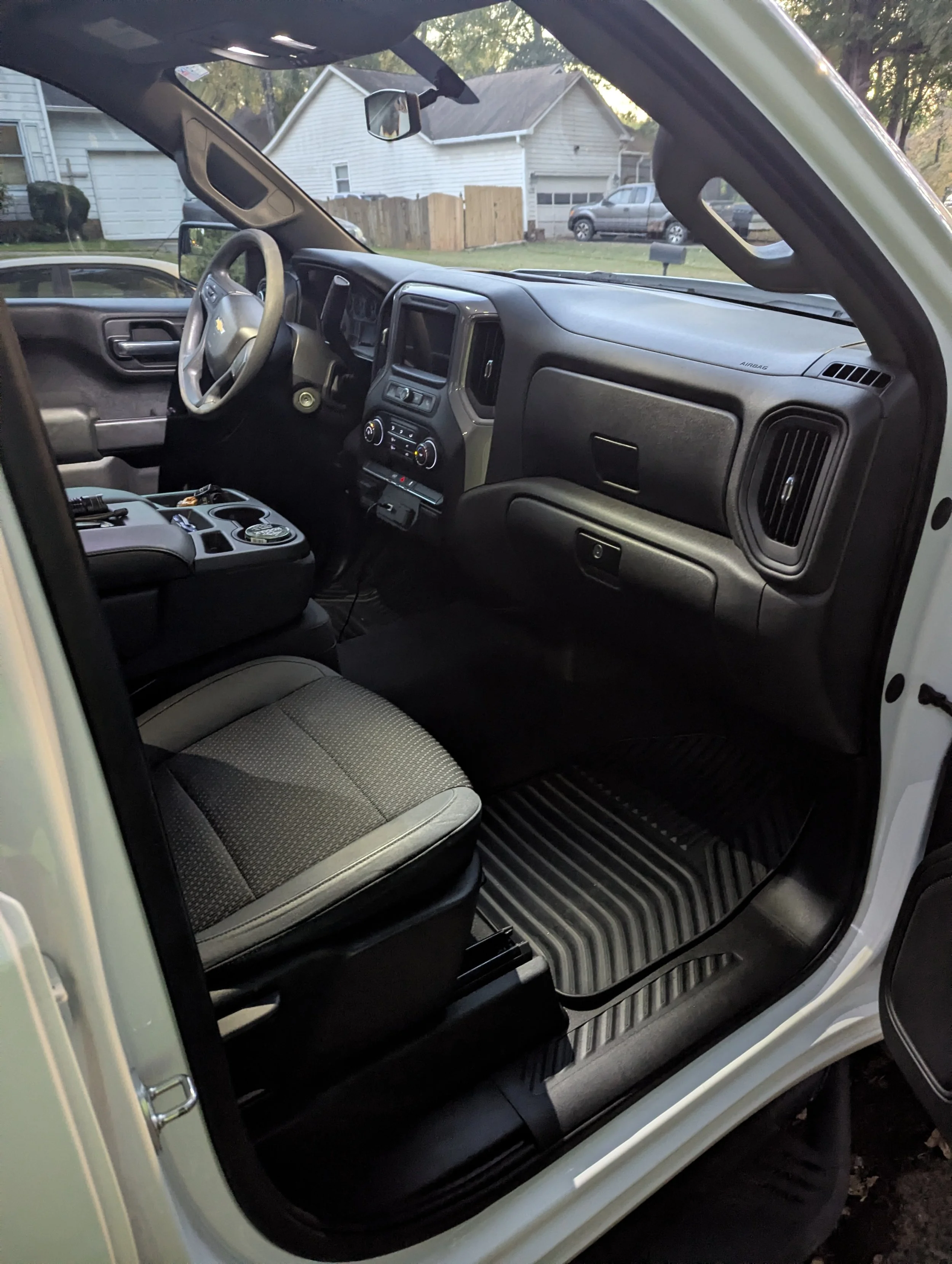 Inside view of a vehicle's front interior, showing the dashboard, steering wheel, driver and passenger seats, and floor mats, with residential houses visible through the windshield.