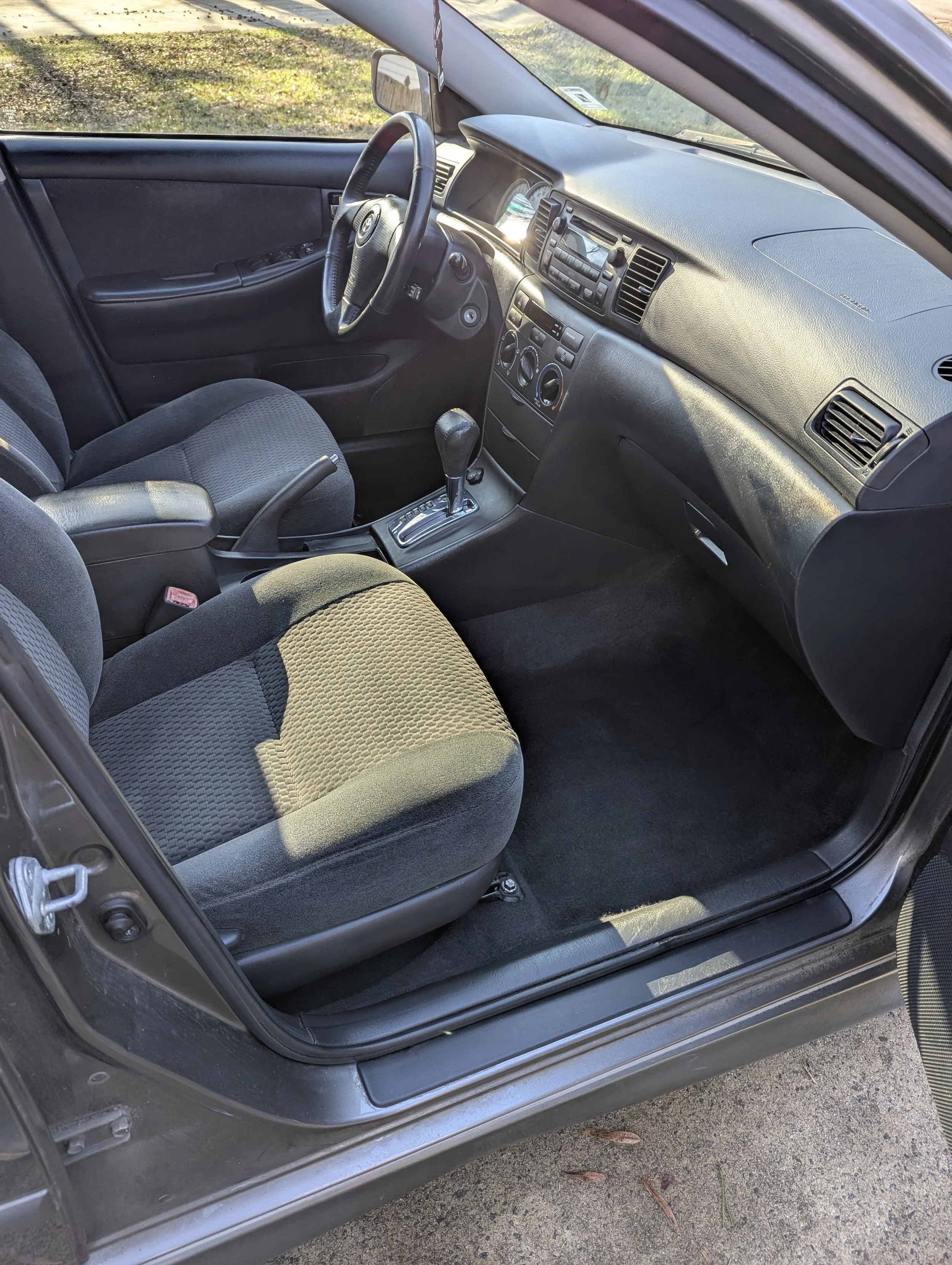 Interior view of a gray car with cloth seats, dashboard, steering wheel, gear shift, and front passenger seat, parked outdoors.