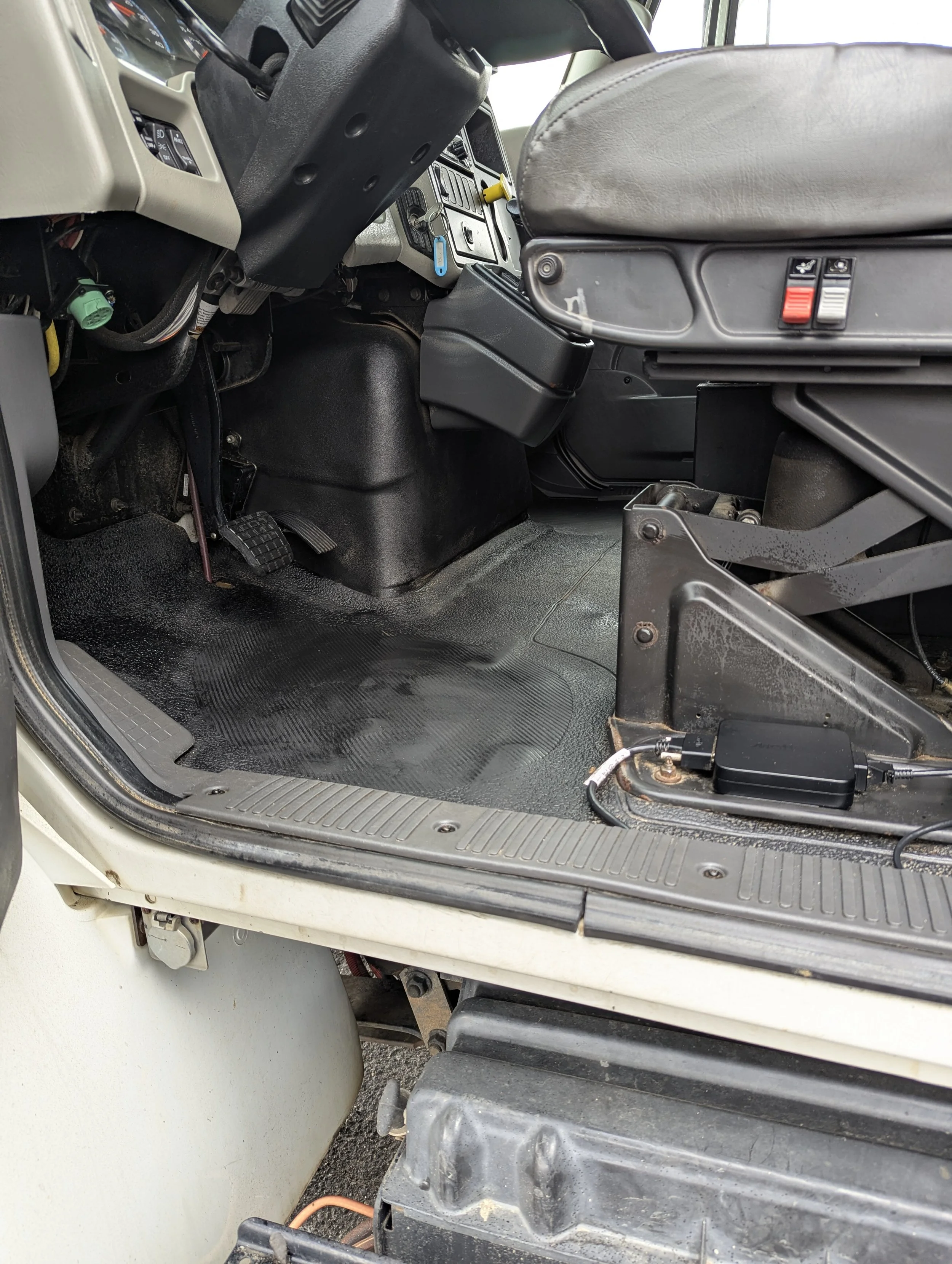 Interior of a vehicle's driver's side, showing the dashboard, steering column, seat, and floor area with some electronic wiring and components.