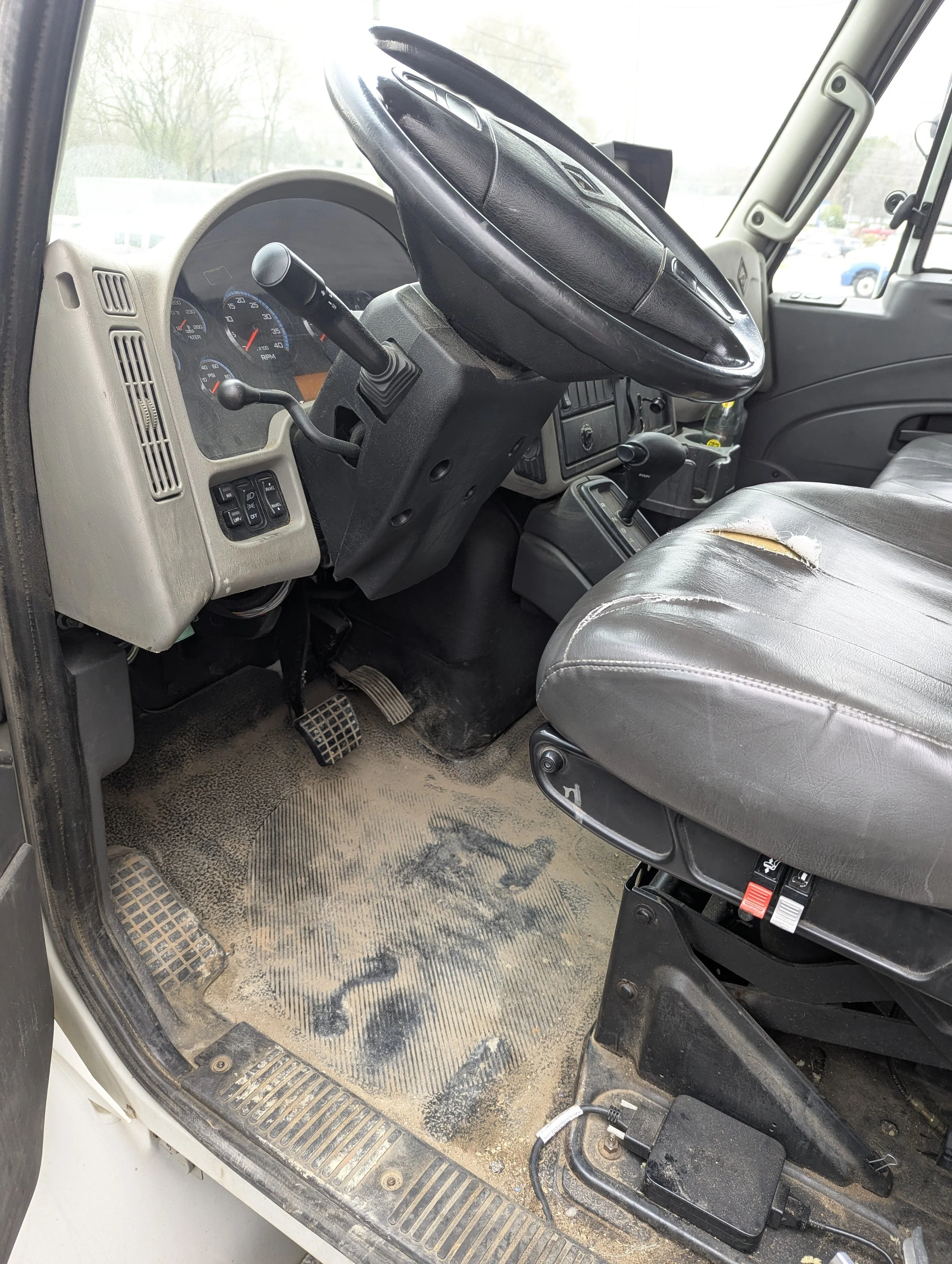 The image shows the interior of a dirty vehicle cab with worn-out flooring, a steering wheel, speedometer, and gear shift, indicating heavy use.