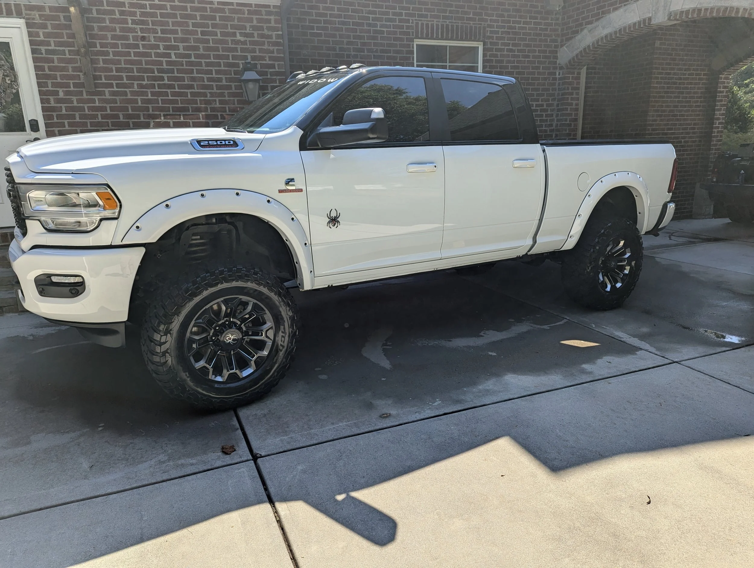 White pickup truck with black wheel arches, off-road tires, black custom wheels, and spider decal on the side, parked on a driveway in front of a brick house.