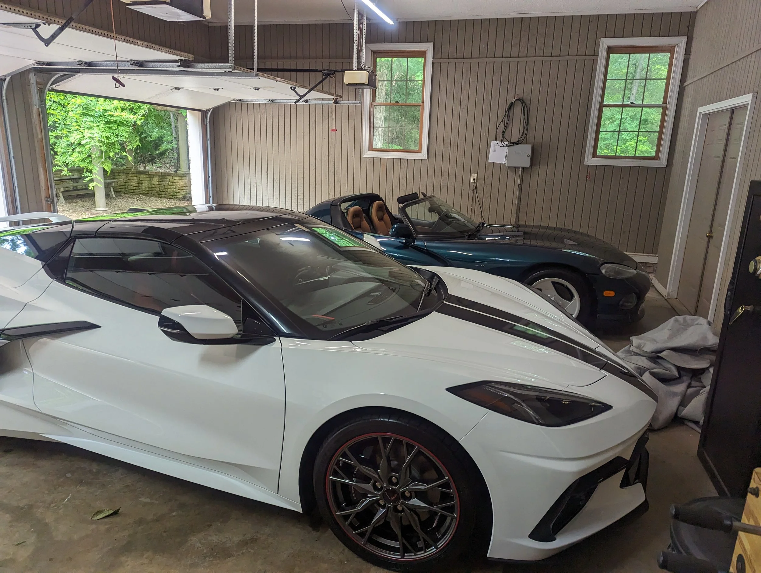 A garage with a white sports car in the foreground and two other sports cars in the background, beige wooden walls, windows, and an open garage door showing greenery outside.