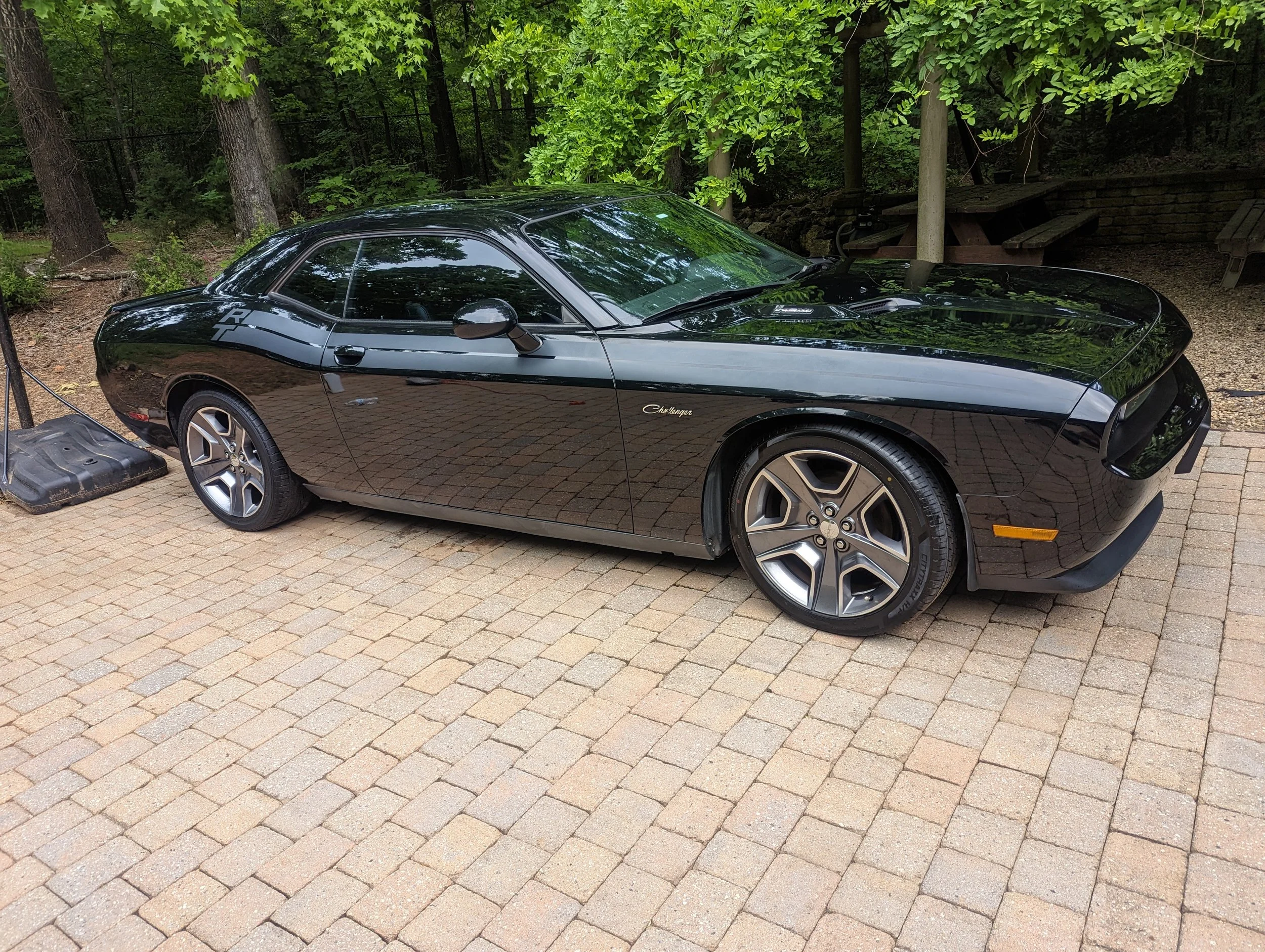 Black Dodge Challenger parked on a brick driveway, surrounded by trees and outdoor seating.