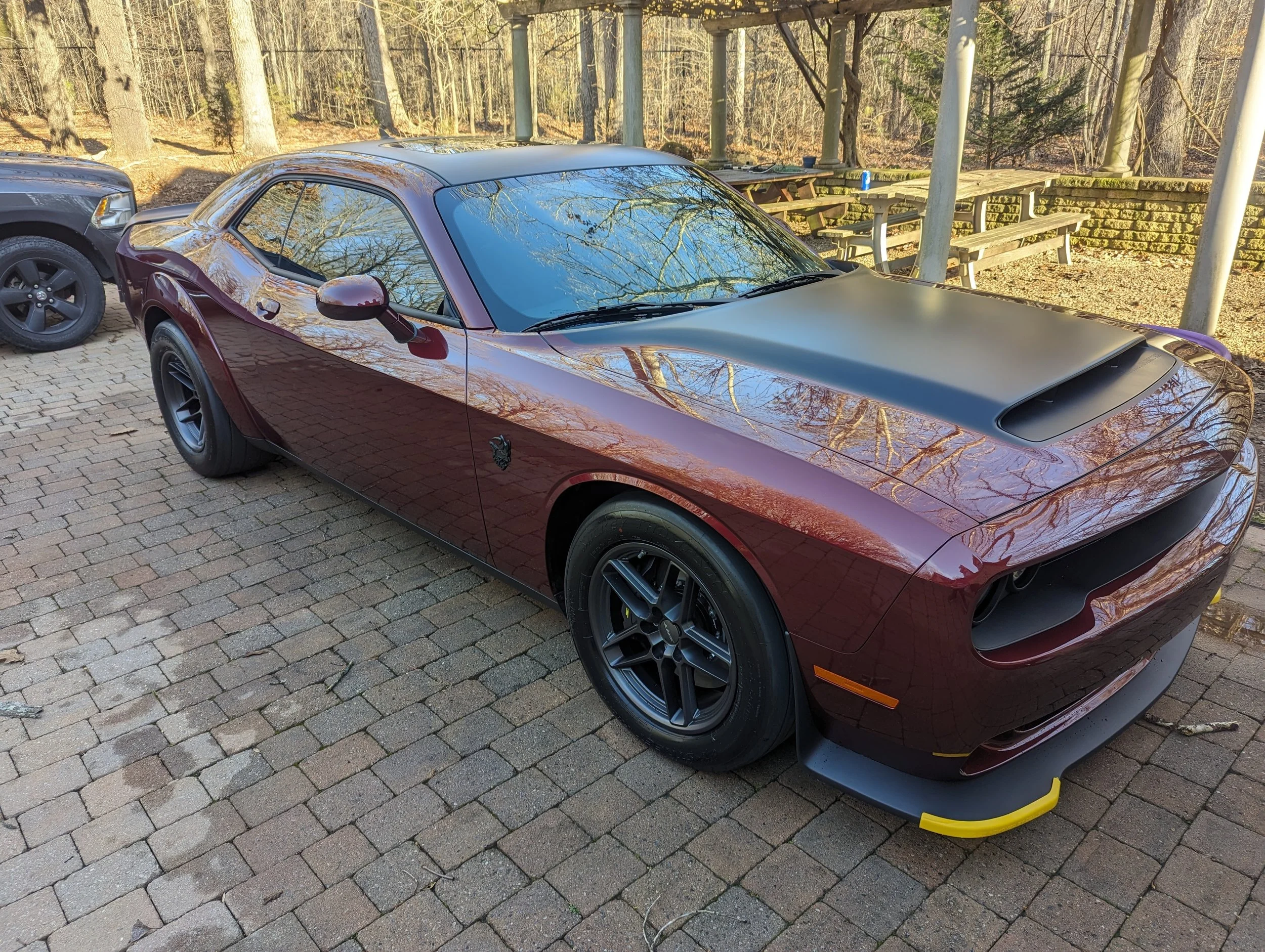 A maroon sports car with black wheels parked on a brick pavement next to a black SUV, outdoors with trees and a wooden picnic area in the background.