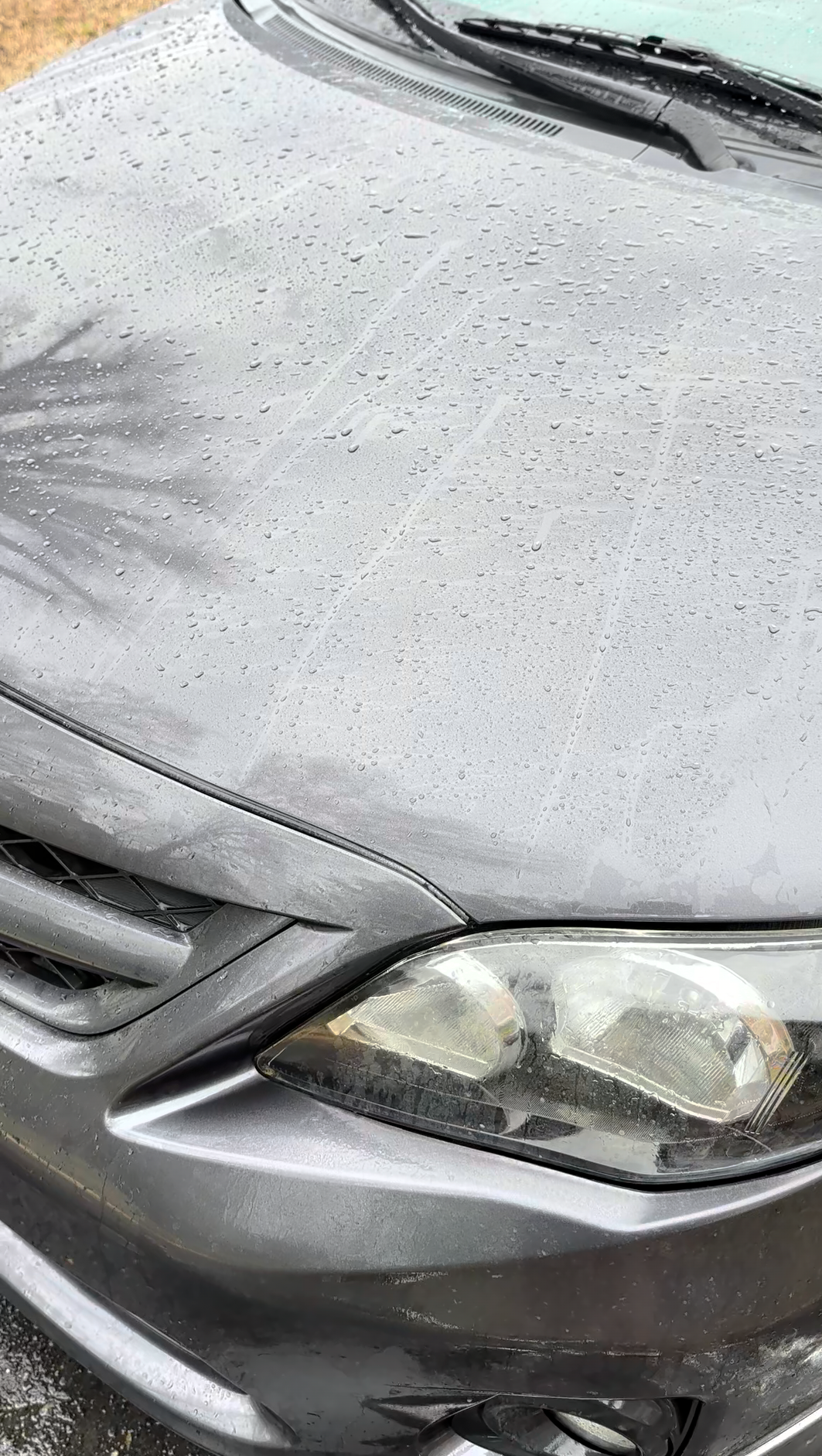 Close-up of a silver car's front end with rain droplets on the hood and windshield, showing the front headlight and part of the grill.