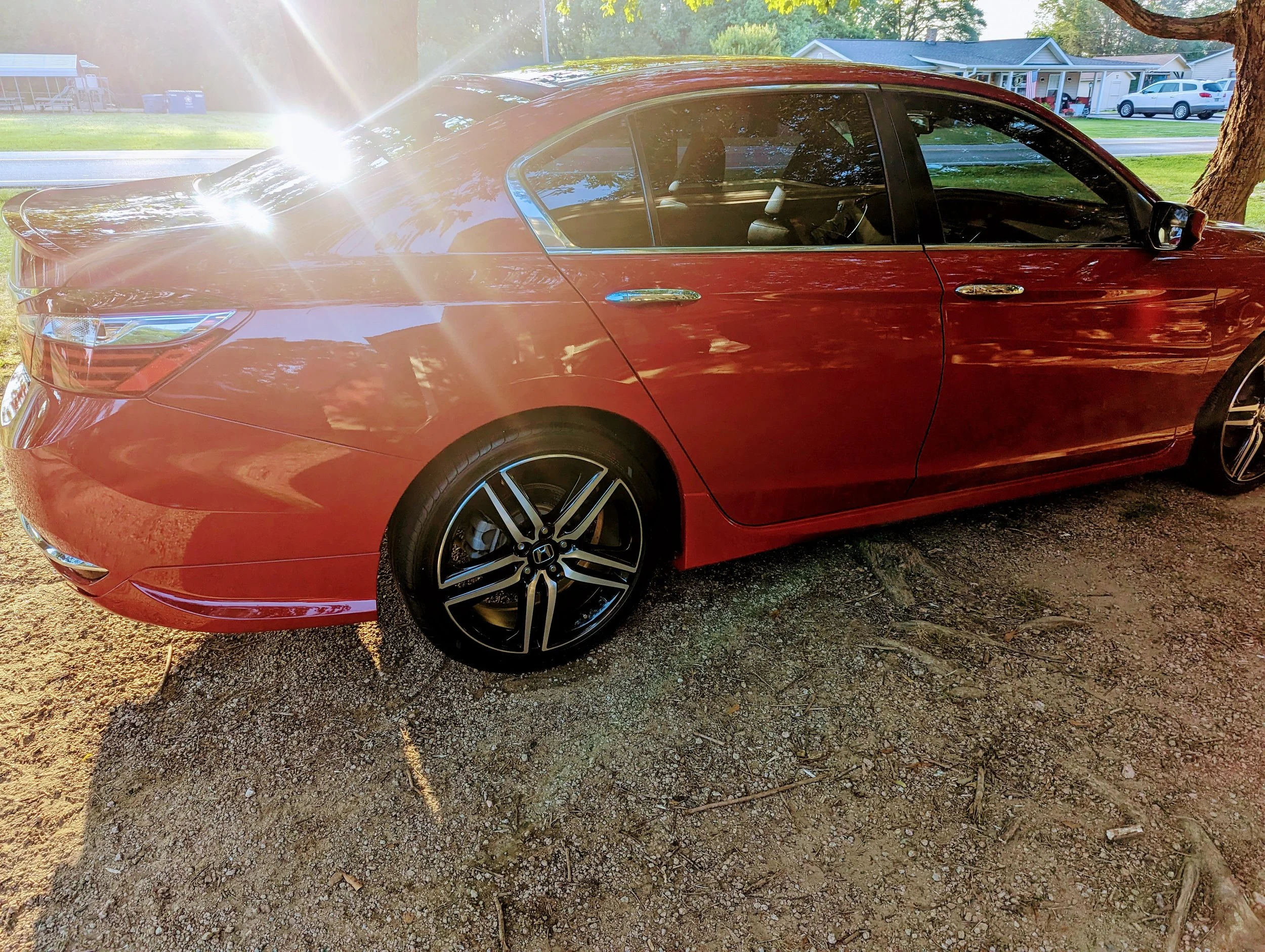 Red sedan parked on a dirt ground with trees and houses in the background, sunlight causing glare on the car.