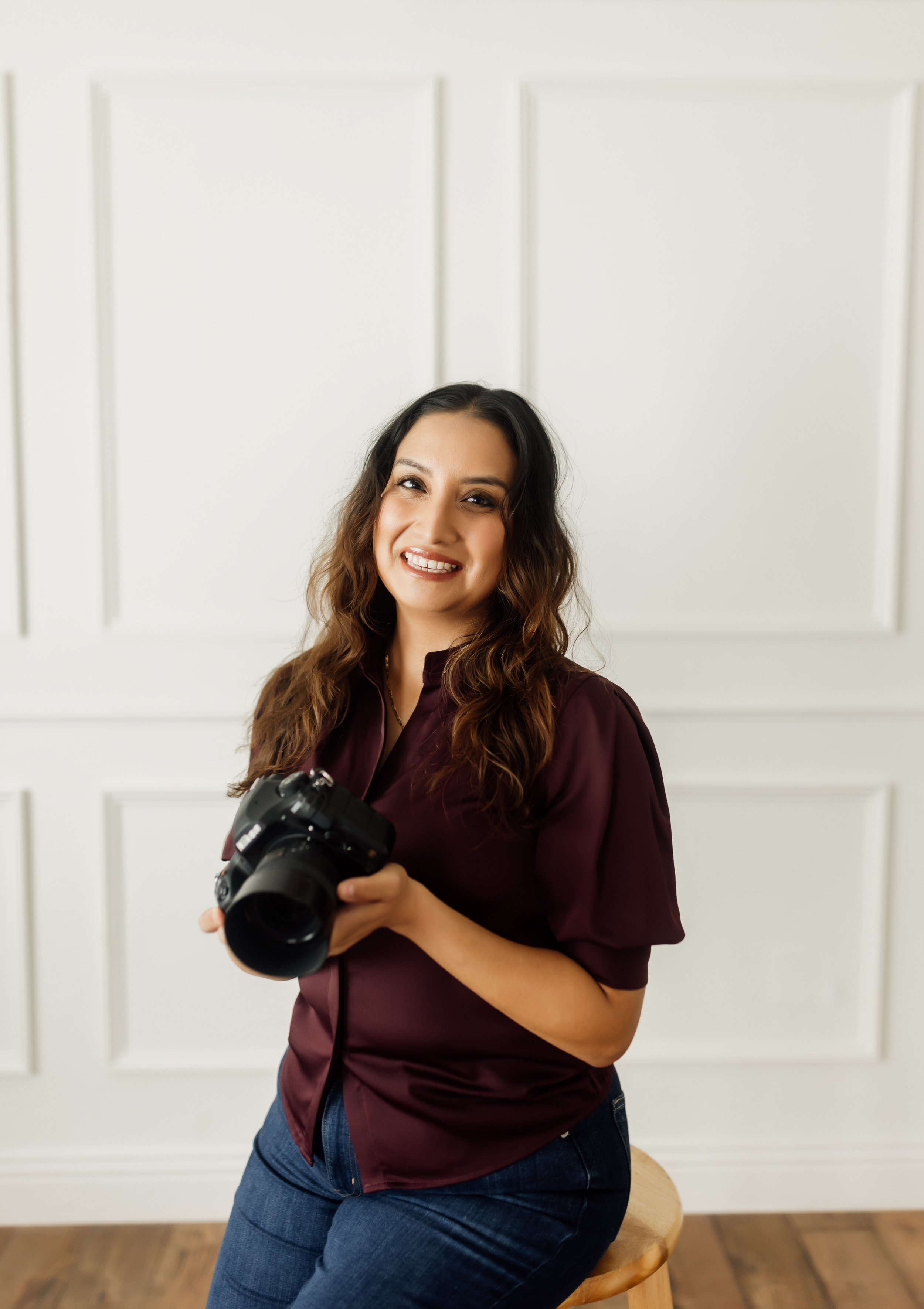 A woman with long wavy brown hair smiling and holding a DSLR camera, sitting on a stool in a room with white paneled walls and wooden floor.