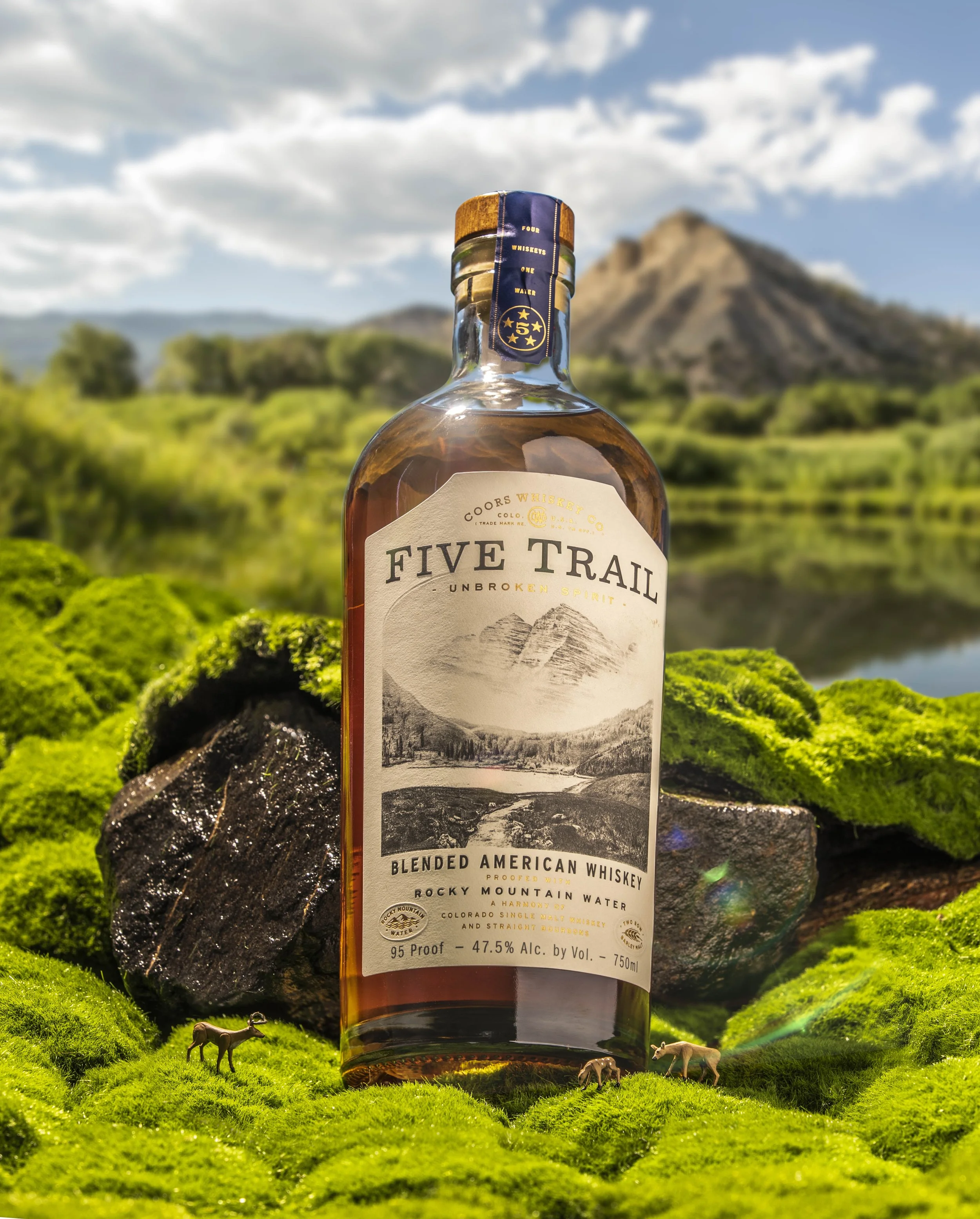 Bottle of Five Trail blended American whiskey with a mountain landscape label, set among green moss and rocks with small deer figurines at the base, and a mountain and cloudy sky in the background.
