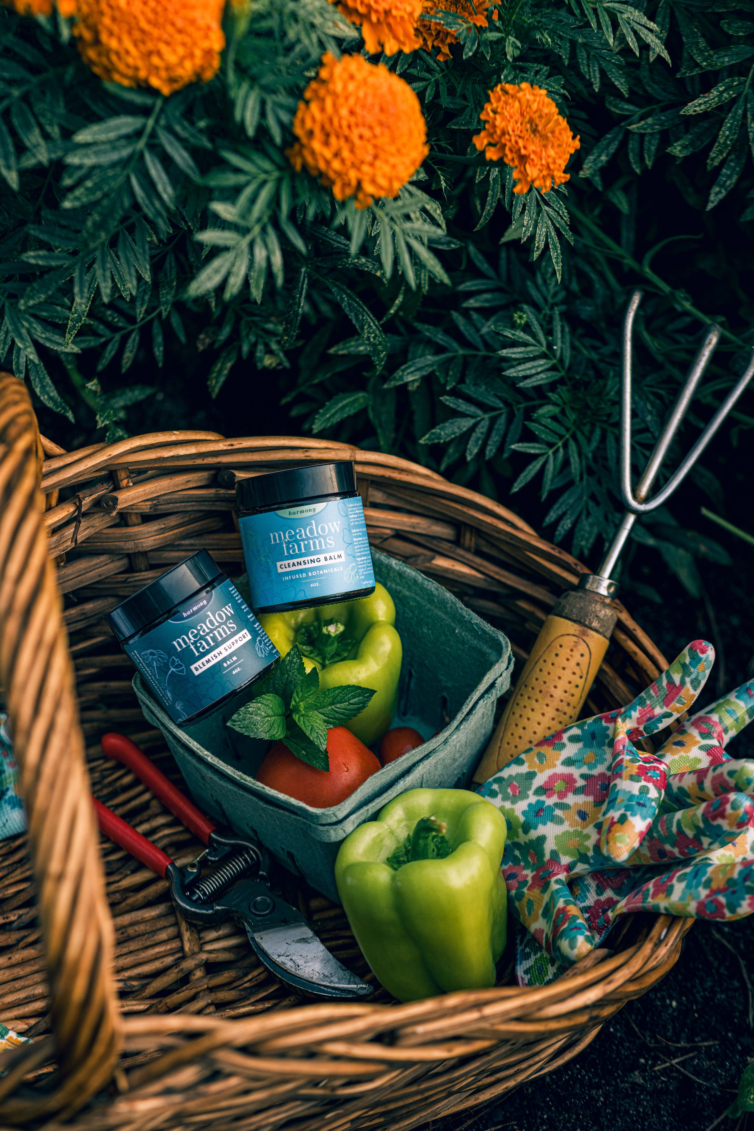 A wicker basket contains green bell peppers, a red tomato, and a yellow apple, with gardening tools like a pruning shear and a gardening fork nearby. There are also skincare products labeled 'Meadow Farms' placed inside the basket, and a colorful cloth is draped over the edge of the basket. Bright orange marigolds and green foliage are in the background.