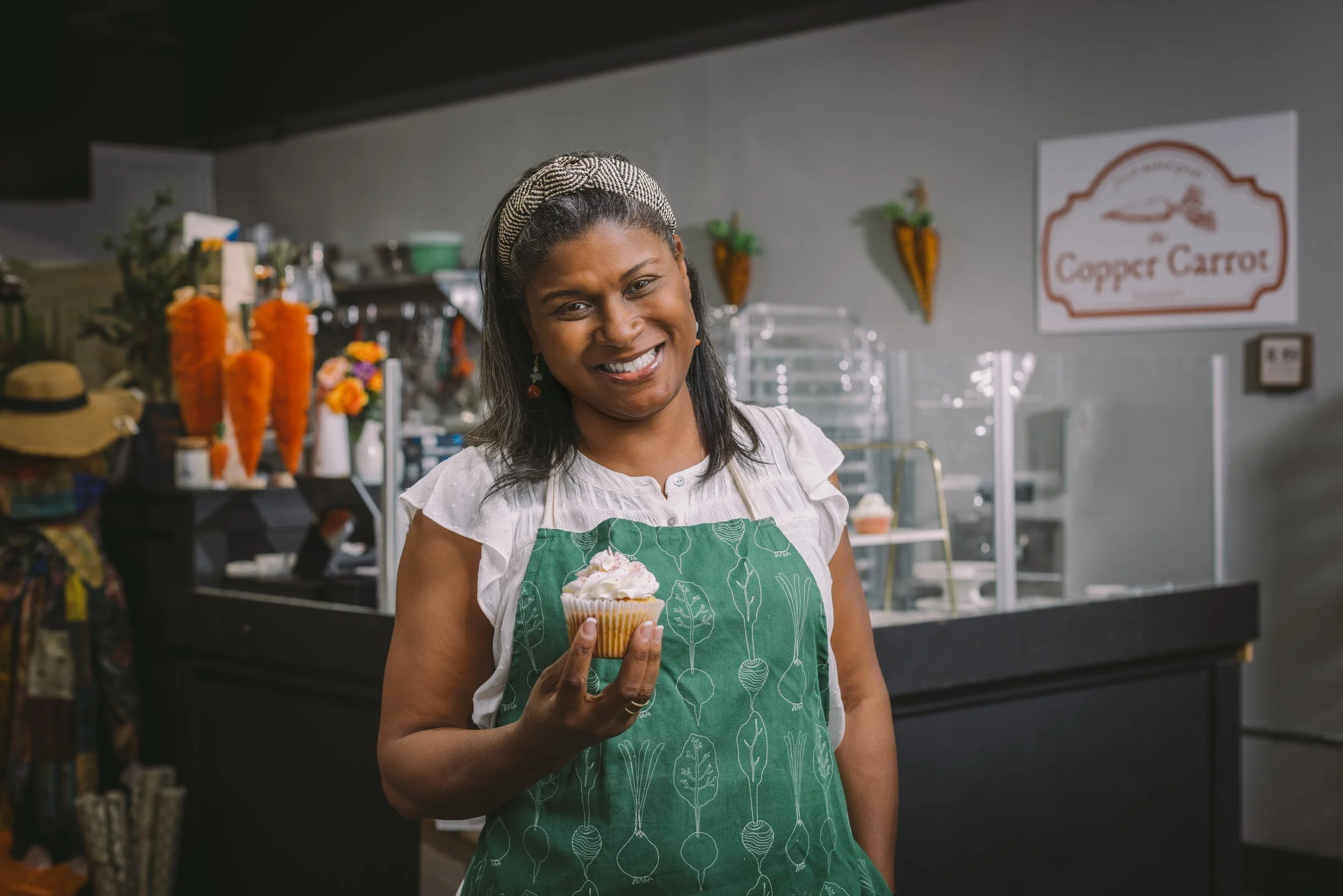 Smiling woman holding a cupcake in a bakery with a sign that reads 'Copper Carrot'.