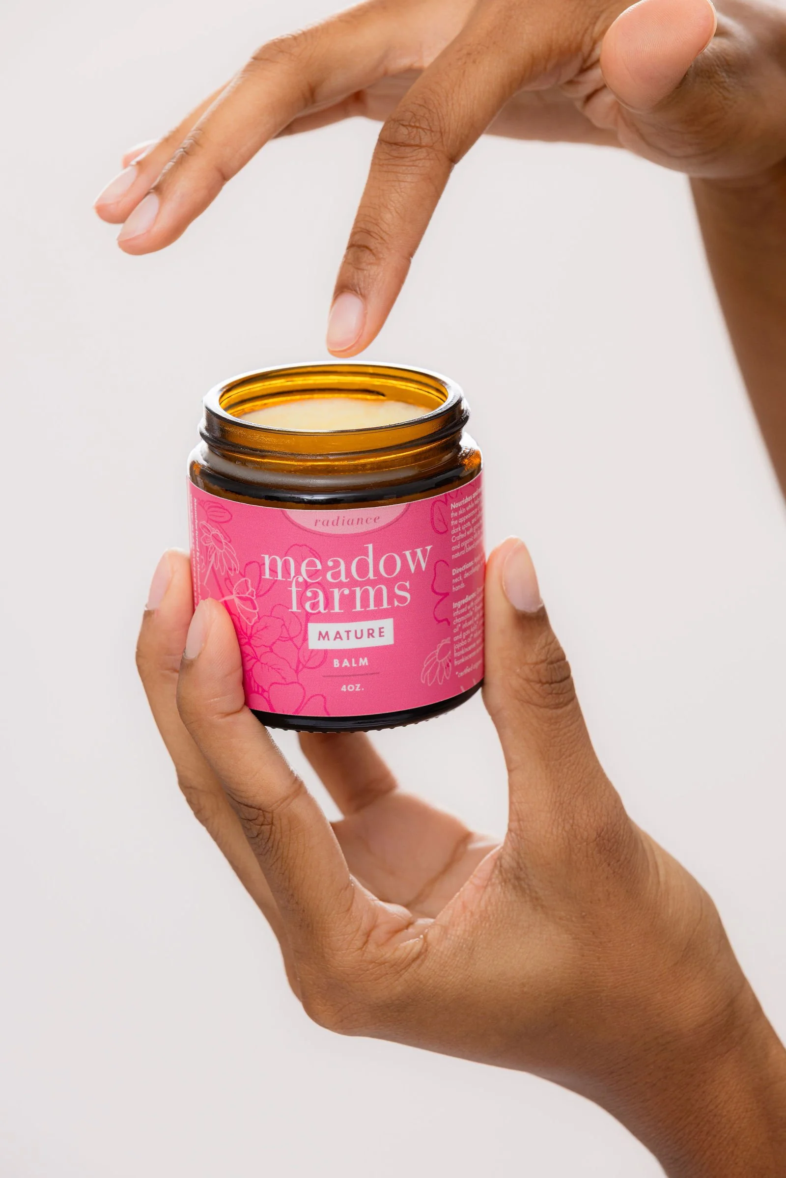 Person holding a pink jar of Meadow Farm's Mature Balm with two fingers, with another hand dipping a finger into the balm.
