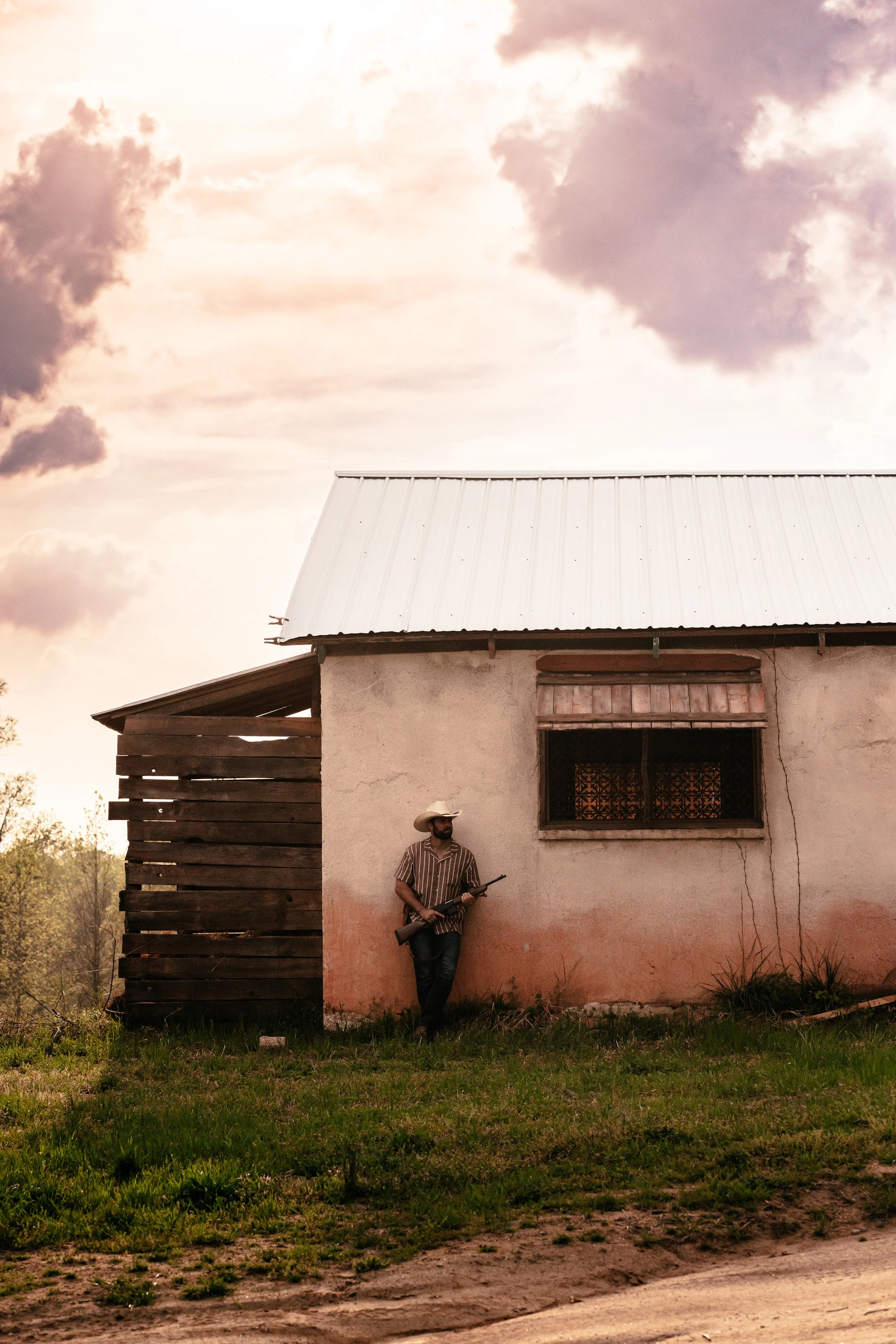 Man wearing cowboy hat and striped shirt leaning against a rustic house wall, holding a rifle, under a cloudy sky, in a rural setting.