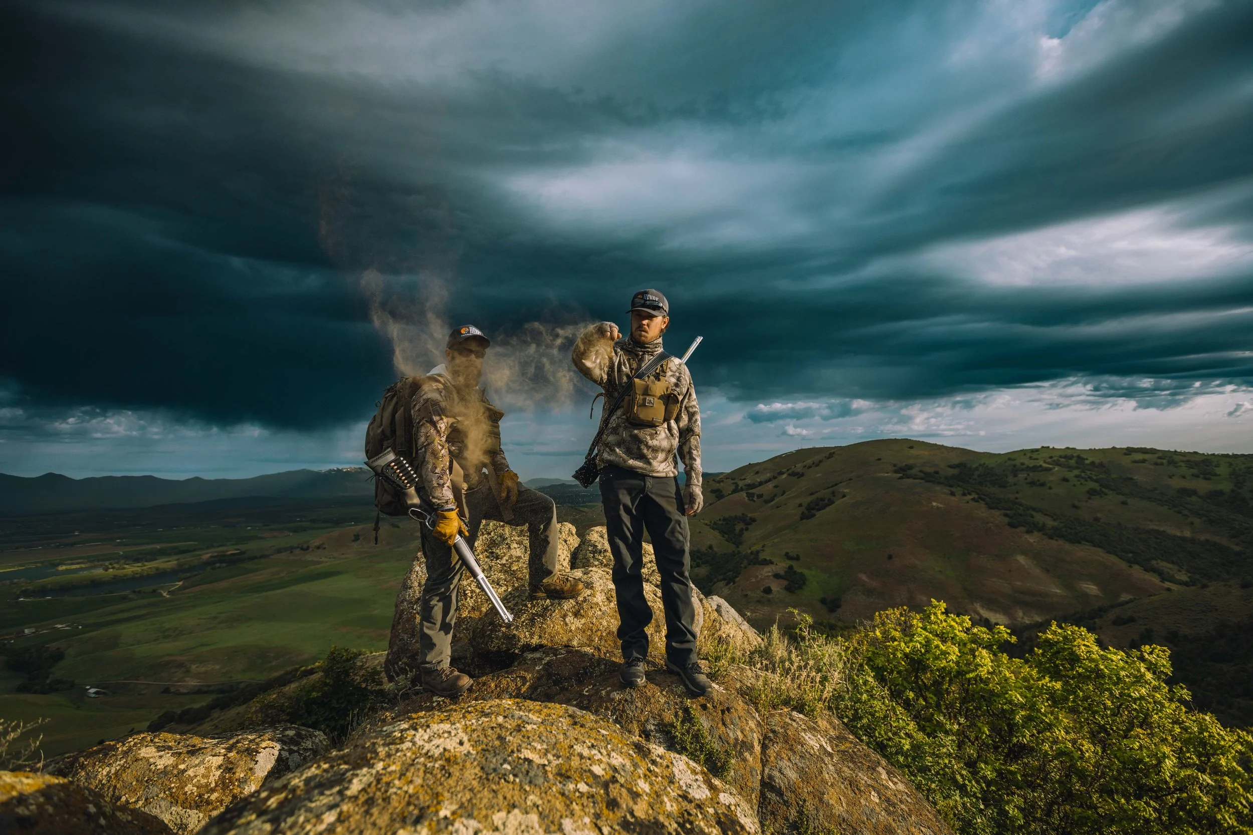 Two hunters standing on rocky hilltops with a dramatic cloudy sky and rolling green landscape in the background.