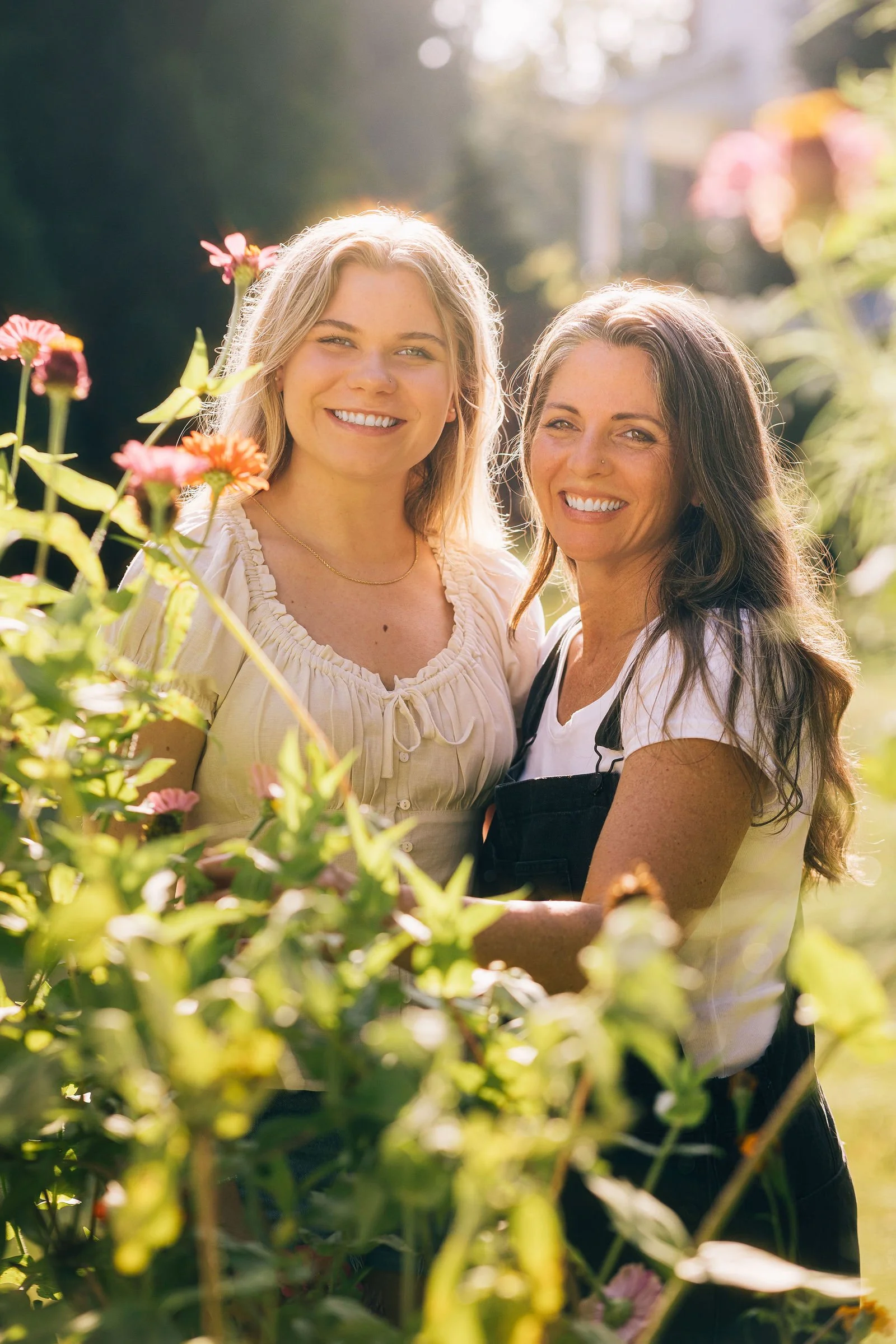 Two women smiling outdoors in a garden with pink flowers and greenery, sunlight shining through the trees.