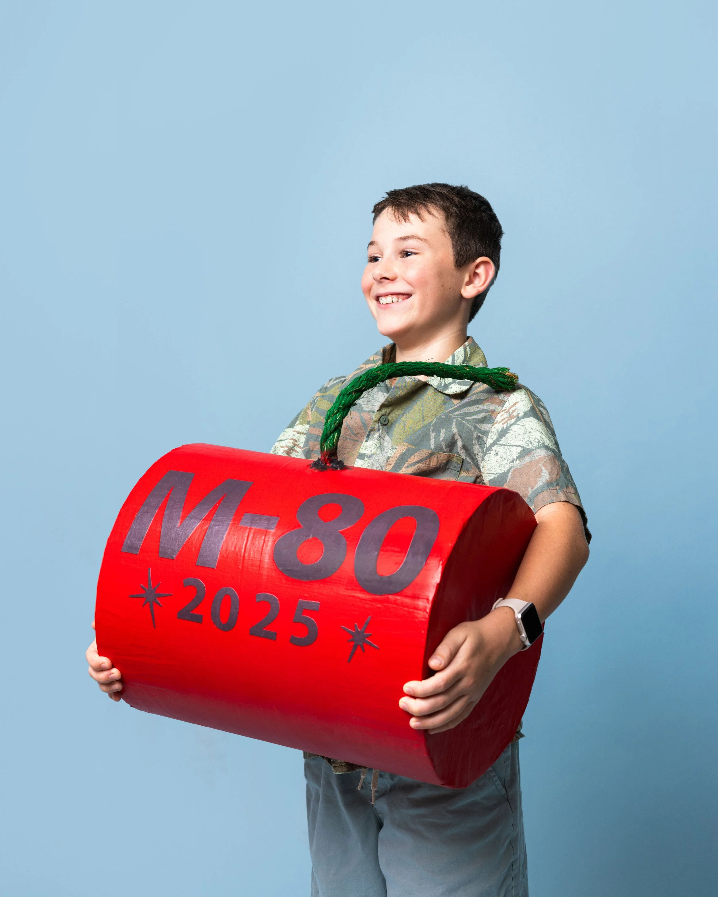 A smiling boy in a camouflage shirt holding a large, red, cylindrical mock bomb with 'M-80 2025' written on it, against a plain blue background.