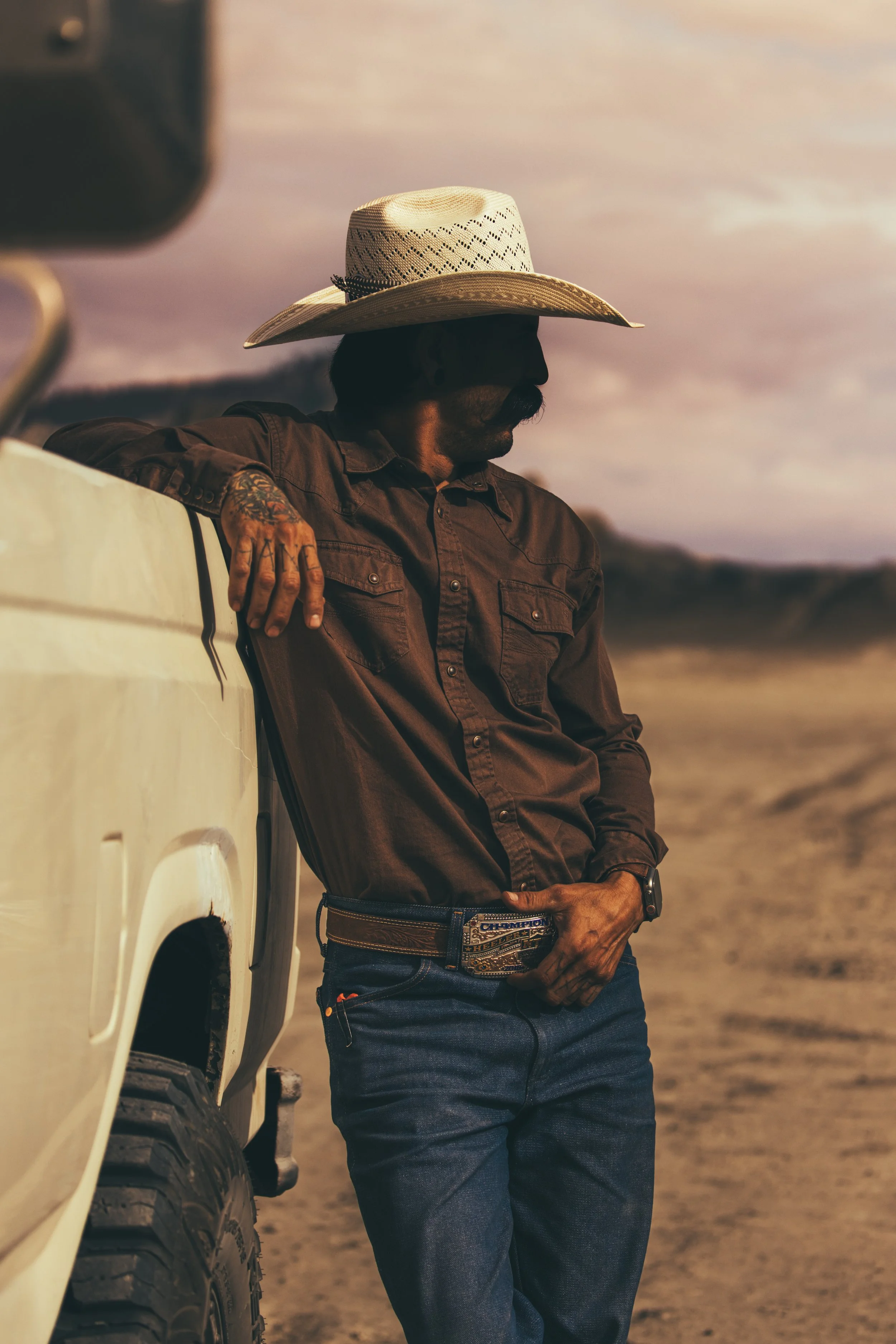 A man wearing a large cowboy hat, dark brown shirt, and jeans standing outdoors near a cream-colored vehicle during sunset.