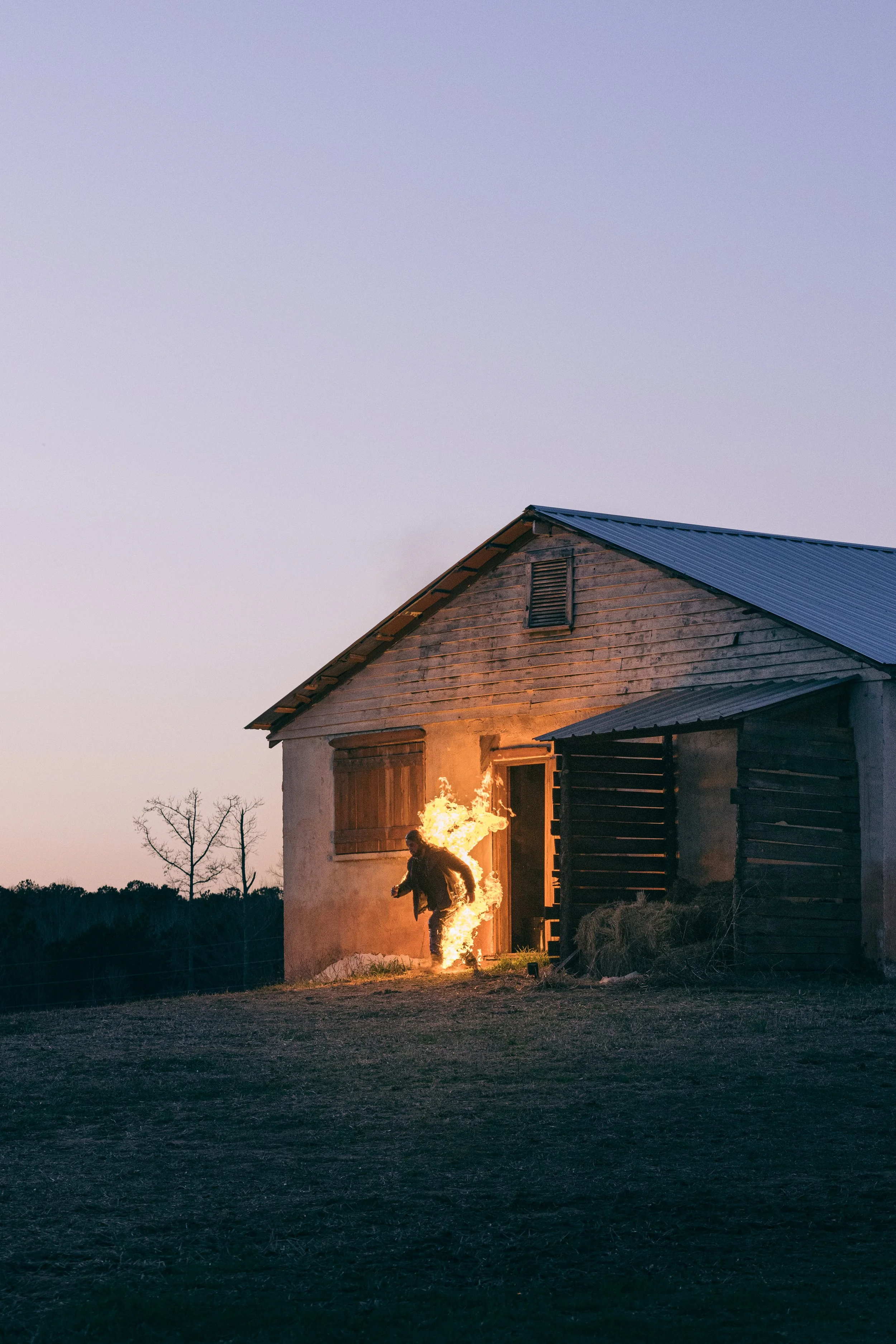 A person running out of a house on fire during dusk or dawn in a rural area with trees in the background.