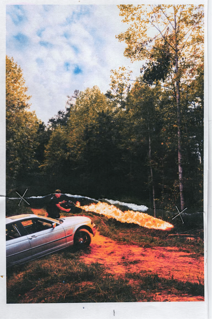 A person standing outside in front of a silver sports car, shooting a fire stream into a forested area with yellow and orange autumn trees under a blue sky with some clouds.