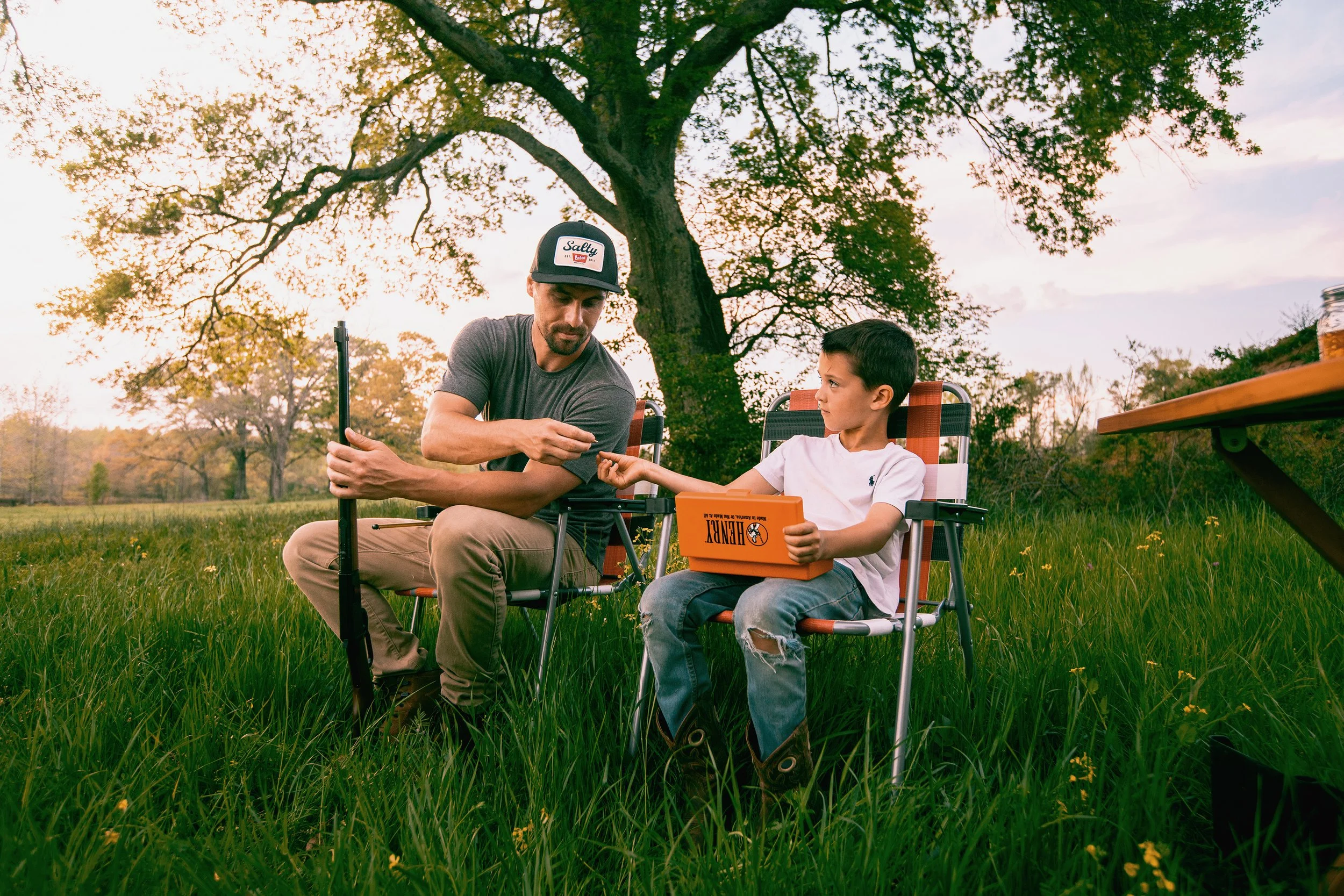 A man and a young boy sitting on lawn chairs in a grassy field under a large tree in the late afternoon. The man is wearing a gray t-shirt and a baseball cap, while the boy is in a white t-shirt and ripped jeans. The boy is holding a toolbox labeled
