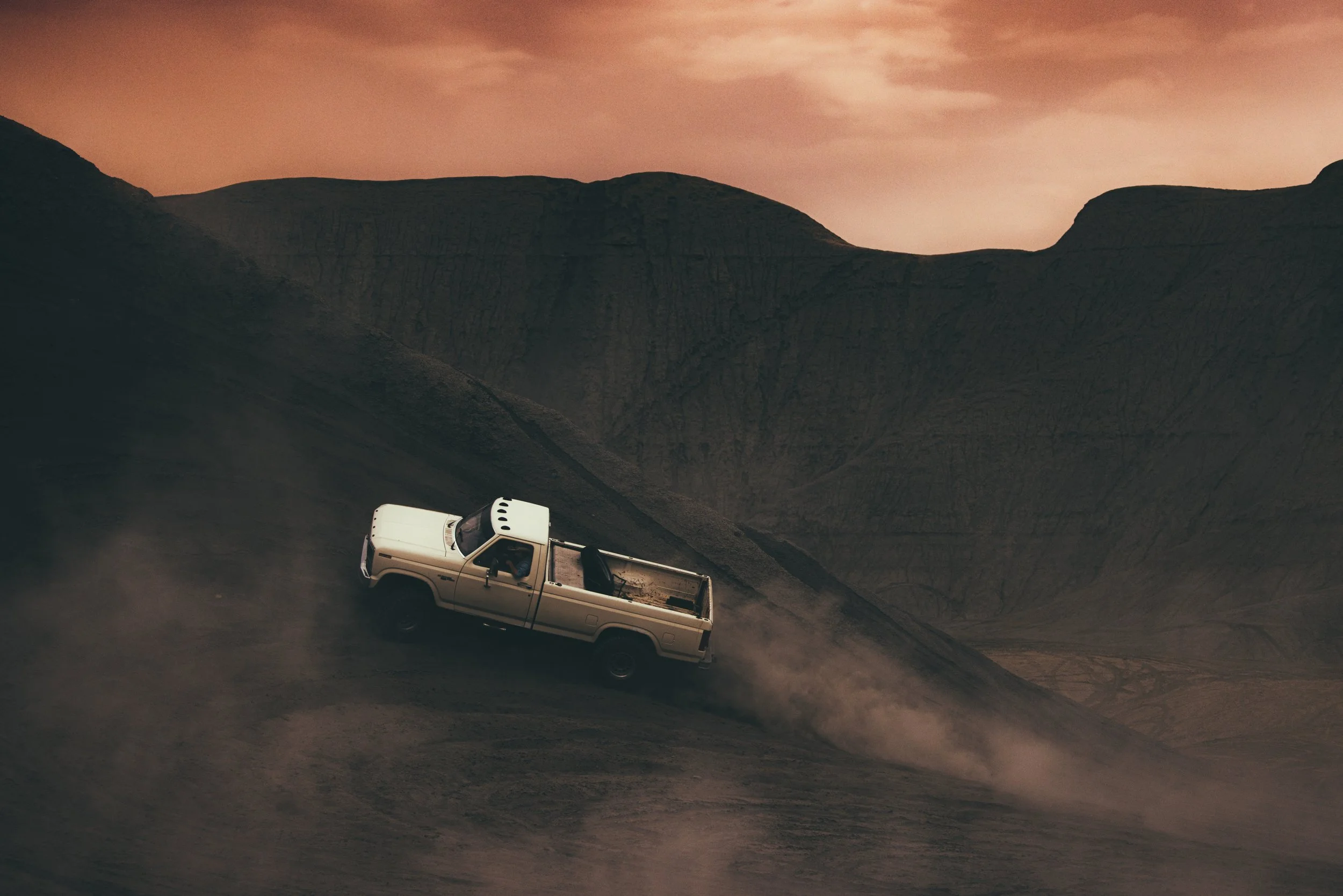 A white pickup truck driving up a dusty slope in a barren, mountainous landscape during sunset with orange hues in the sky.