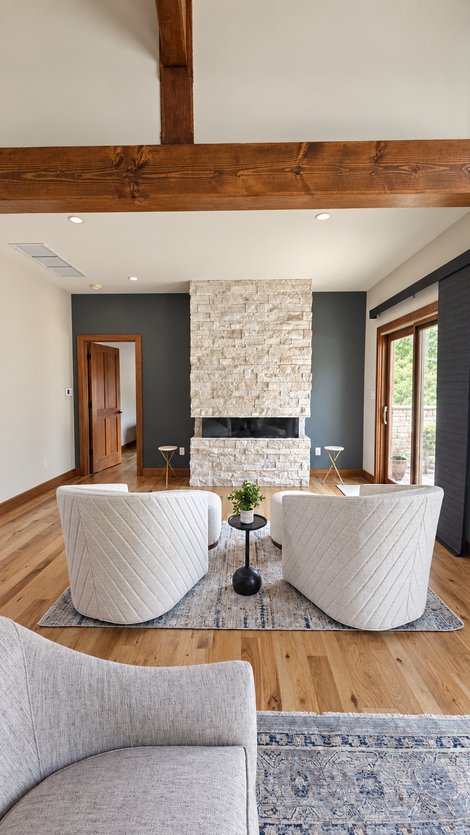 Living room with a stone fireplace, two white armchairs, a small black side table with a plant, hardwood floors, a sliding glass door, and a ceiling beam.