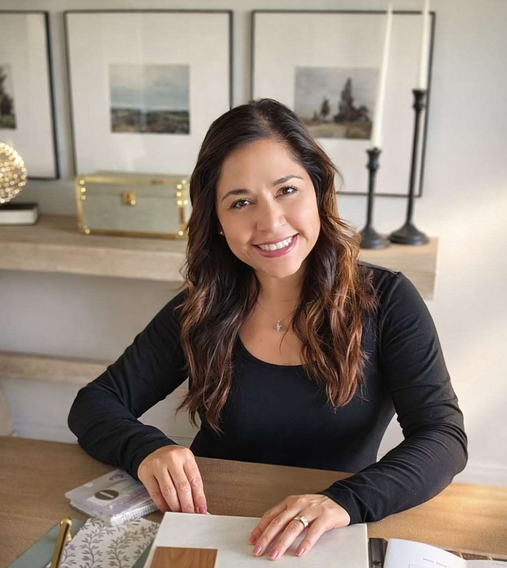 A woman with long brown wavy hair sitting at a table, smiling at the camera in a home interior with framed pictures, candles, and decorative objects in the background.