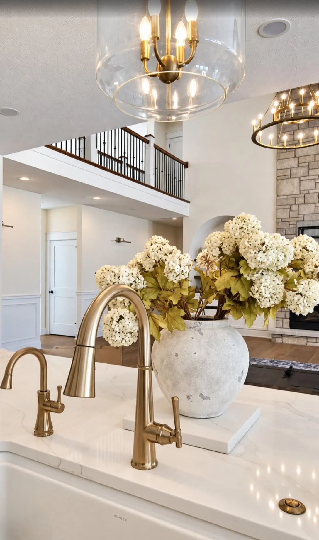 A kitchen with a large white vessel holding white hydrangea flowers, gold faucets, and a view of a living room with a brick fireplace and chandeliers.