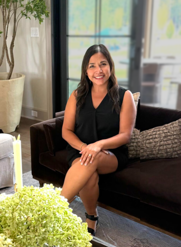 A woman with long dark hair smiling, sitting on a dark brown couch in a bright living room with large windows, a potted plant, a gray pillow, and a table with a flower arrangement in front of her.