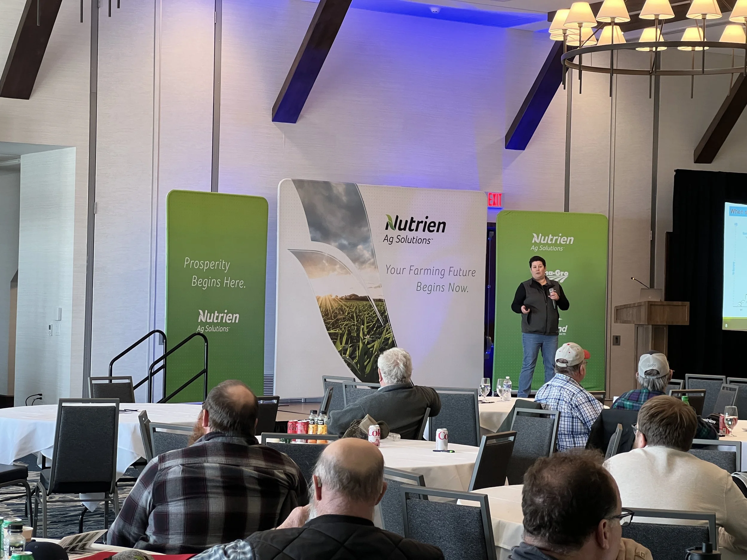 A speaker presenting at a Nutrien Ag Solutions conference with audience members seated at tables in a large conference room.